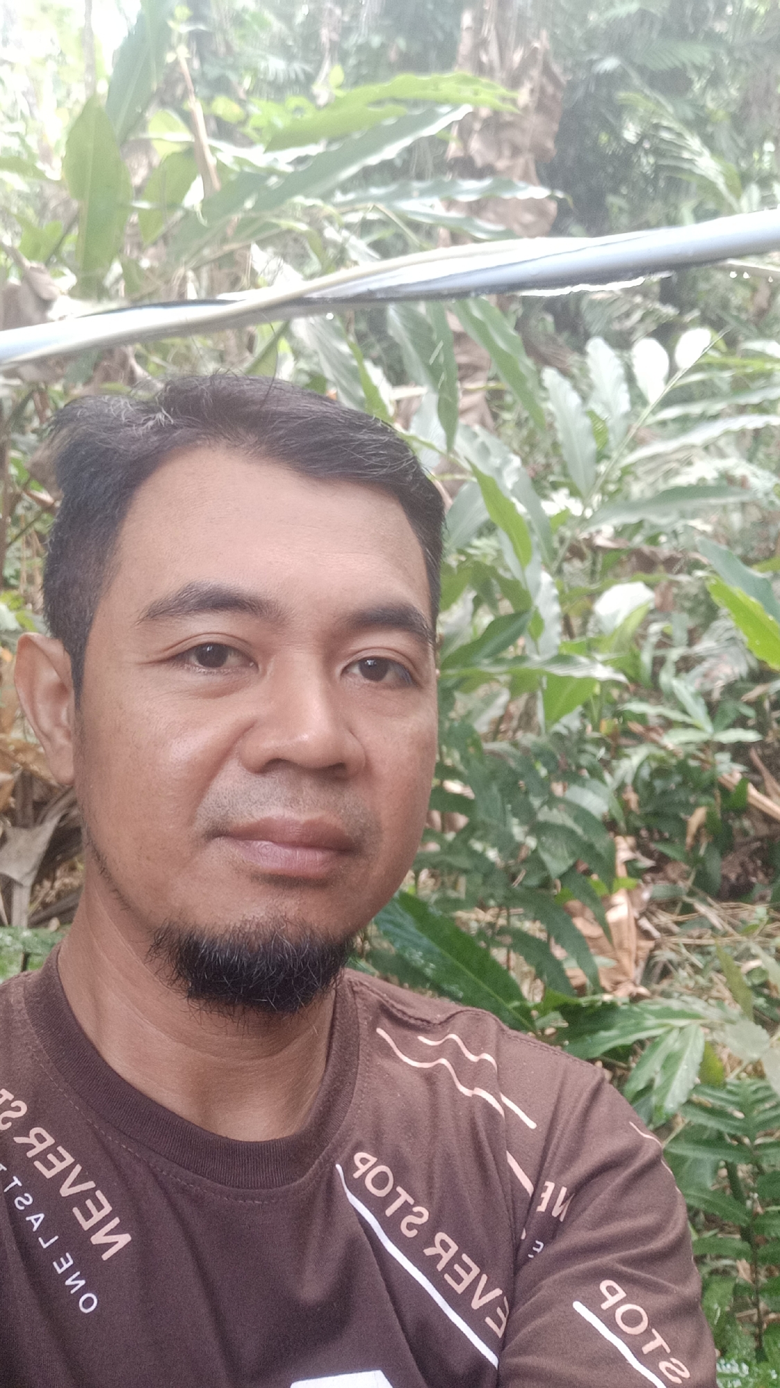 A man with dark hair, a goatee, and brown skin is pictured in a close-up selfie. He is wearing a brown t-shirt with white text that appears to be in a foreign language. The background is lush and green with foliage and trees, suggesting an outdoor, possibly rural setting. The image was taken in Desa Burujul Jaya, Indonesia, as indicated by the metadata. The man's expression is neutral, and he is looking directly at the camera. There are no other people or significant objects in the frame.