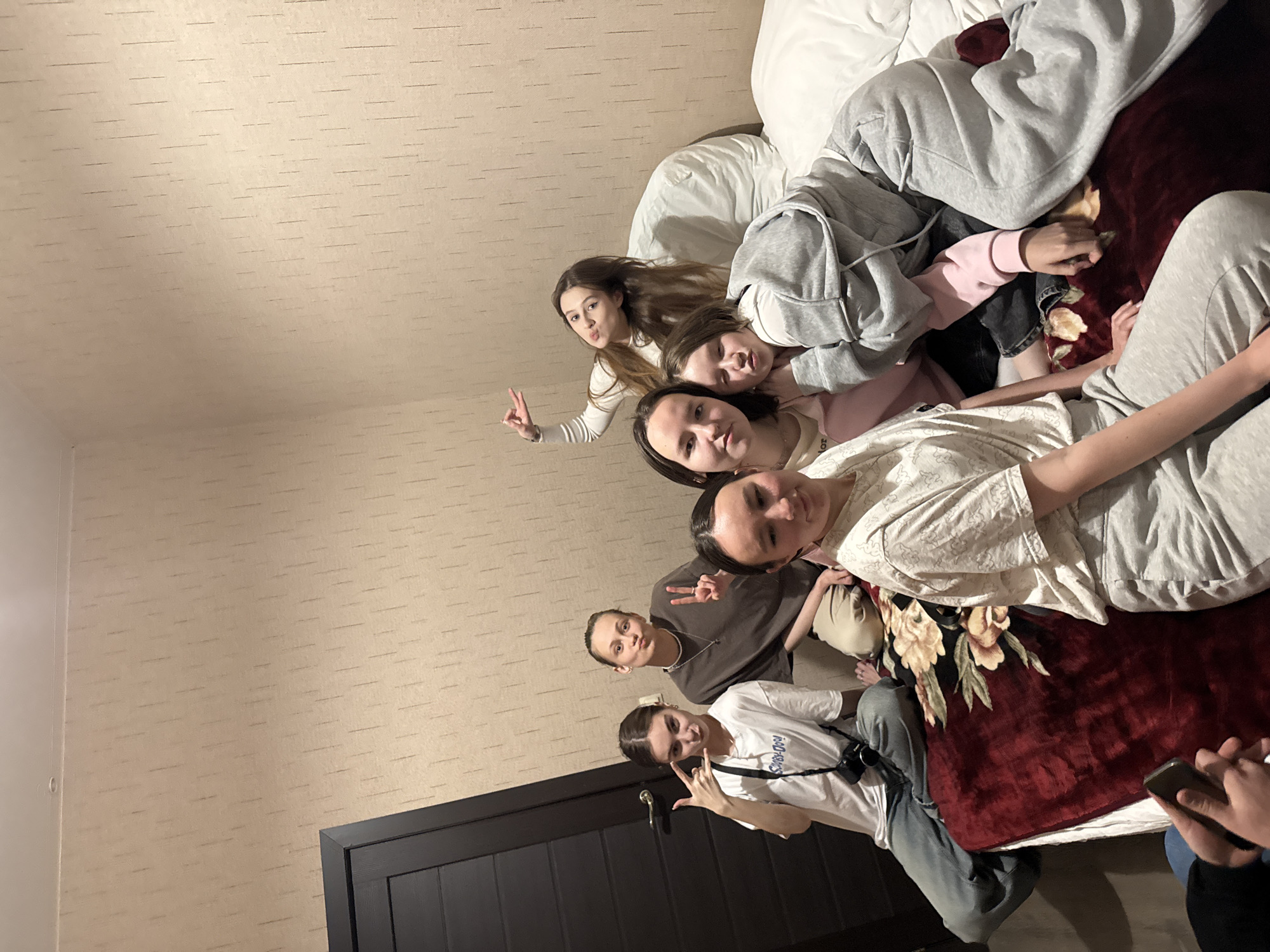 The image appears to be taken indoors, possibly in a private residence, given the wallpaper and door. Seven young women are posing, sitting, and lying on a bed with white bedding and a red patterned blanket. Some are making hand gestures. One woman wears a white t-shirt with a camera. A portion of a cell phone and a hand are in the lower right corner. The location is stated to be in Кашино, Russia.