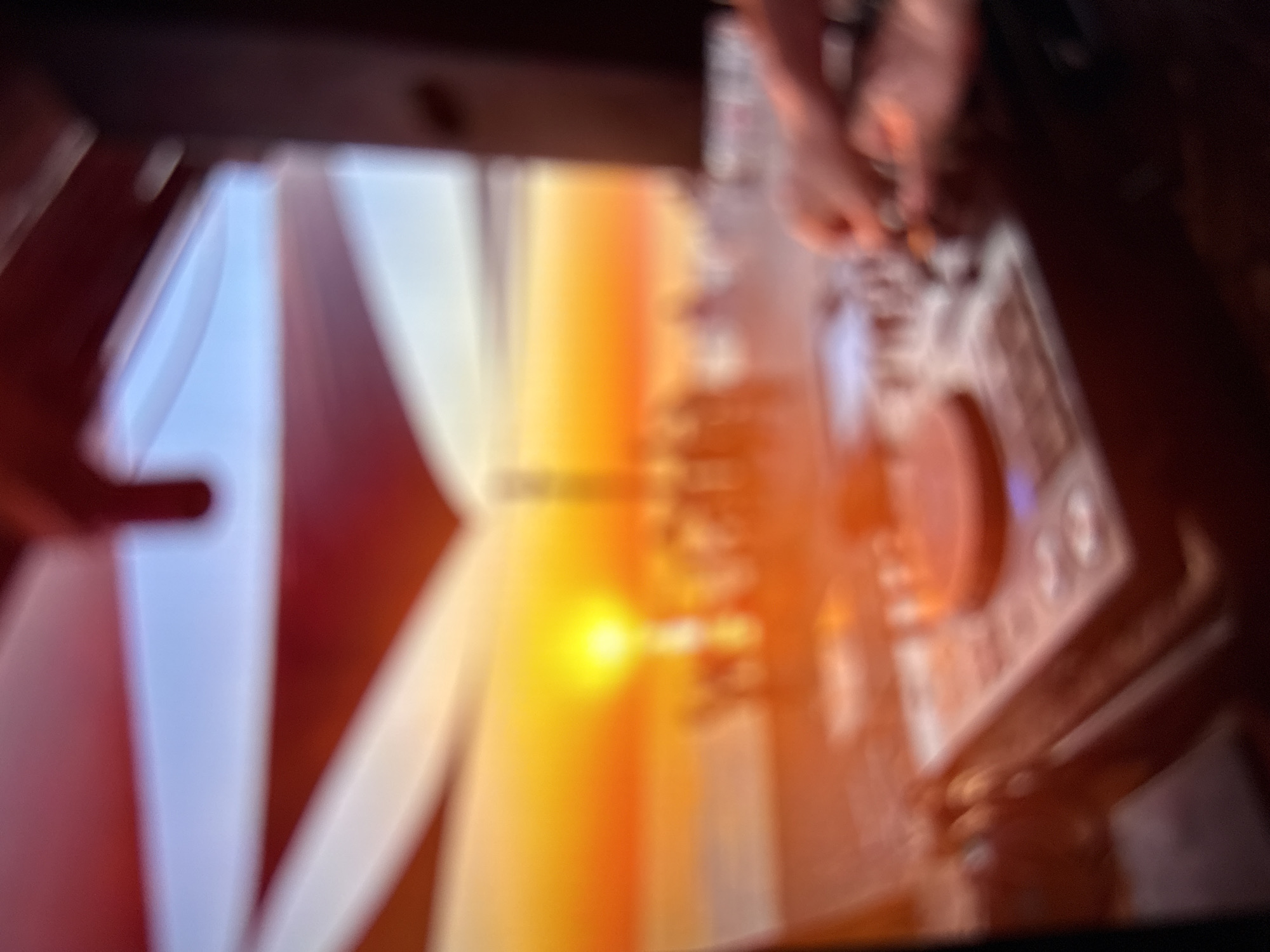 The image displays a heavily blurred outdoor scene at sunset in Kardzhali, Bulgaria. In the immediate foreground, a DJ mixing console is discernible, featuring a prominent circular jog wheel and a cluster of control knobs and buttons. Two blurred hands are positioned over the equipment, actively engaging with the controls on the right side of the console. The mid-ground reveals a vibrant, intensely bright yellow-orange sun low on the horizon, casting a warm glow across the entire environment. Below the horizon, numerous indistinct, silhouetted figures are visible, indicative of a crowd or gathering. A faint, blurred vertical structure rises from the horizon line near the center. The upper portion of the scene shows blurred red and white fabric structures, likely canopies or sails, set against a lighter blue sky. A dark, vertical element frames the right side of the image, possibly a speaker or a structural pillar. The dominant lighting is a rich orange-red, characteristic of a sunset, permeating the entire composition.