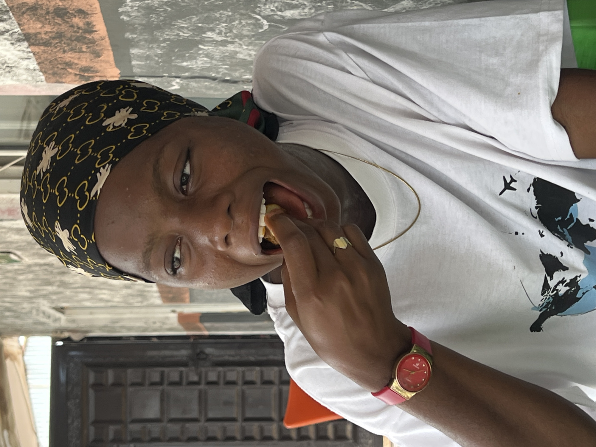 In Nkpor, Nigeria, a young person with dark skin and wearing a black headscarf with a gold pattern is captured in a medium shot, mid-action eating a piece of food. Their mouth is open as they bring the food to their mouth with their right hand, on which they wear a gold ring. A pink-strapped watch with a gold face is visible on their left wrist. The person is dressed in a white t-shirt with a graphic print of a world map and an airplane. The background shows a textured grey wall and a dark metal door. An orange chair is partially visible to the left.
