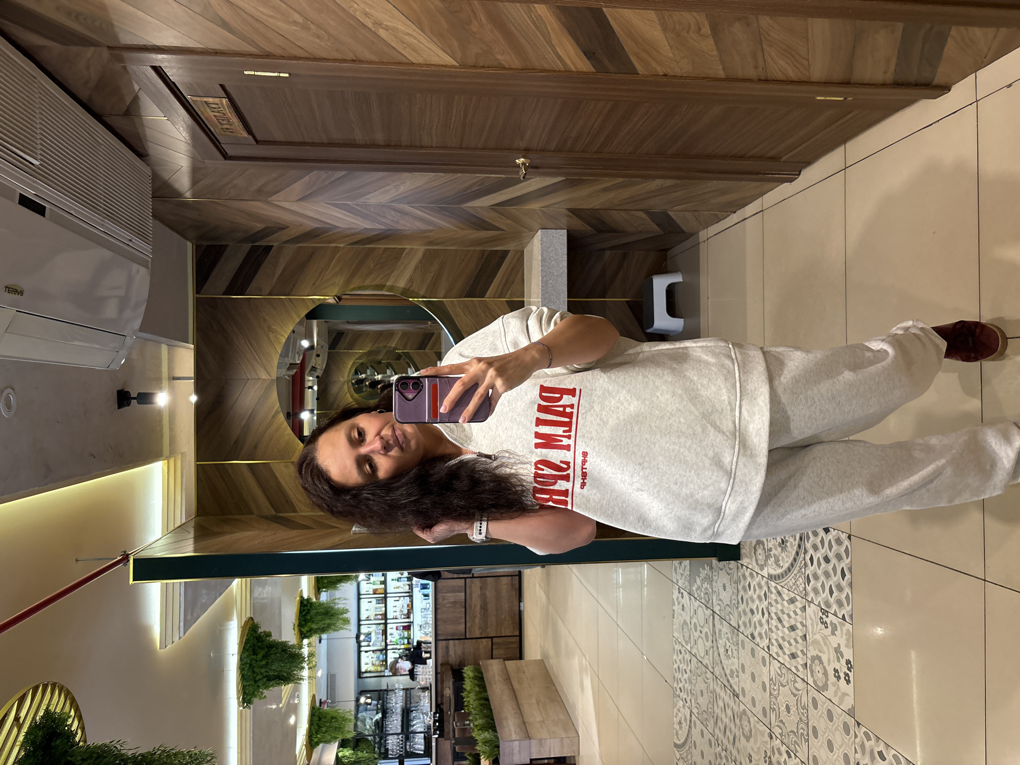 A woman is taking a mirror selfie in what appears to be a restaurant or bar setting. She is holding a smartphone and posing, showing off her hair. She is wearing a light gray sweatshirt and matching sweatpants. In the mirror's reflection, one can see shelves filled with bottles (likely alcoholic beverages), potted plants, and tables. The interior design includes wood paneling on the walls, patterned tiles on the floor near the mirror, and possibly a seating area in the background. The door in the background indicates "ТАУЕЛЯ Б", and the air conditioning unit is labeled as "TEHVMS". Based on this information, the photo was taken in Neftekamsk, Russia, at a restaurant or bar called "Тауеля Б".