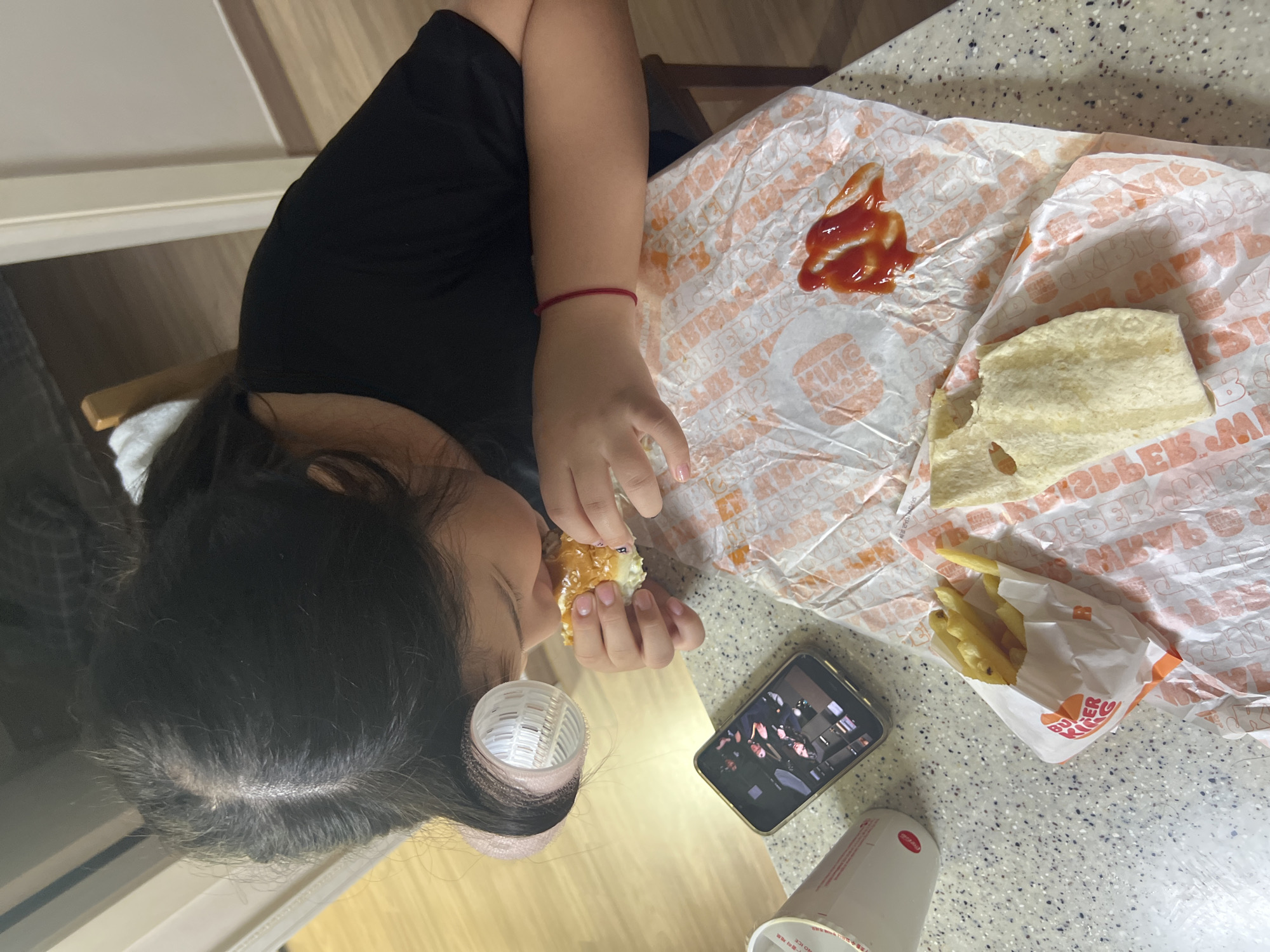 An overhead, rotated shot captures a person eating a fast-food meal at a speckled countertop or table. The person, with dark hair and wearing a black t-shirt, is positioned as if lying down or leaning back, with hair rollers visible in their hair near their ear. They are holding and eating a half-eaten burger, which has sesame seeds on the bun. A red string bracelet is visible on their right wrist, and their fingernails appear to have light nail polish.
On the surface, to the person's right, is white Burger King wrapping paper. A puddle of ketchup is visible on the wrapper. Next to the wrapper, a piece of flatbread or an additional bun part is also present. Further to the right, a small pouch of french fries, branded "BURGER KING," sits on the wrapper. A smartphone, displaying a video or image of multiple people, rests on the table, likely being watched by the person. A white cup, possibly a drink container, is also on the table, with some unreadable text and "NO ICE" visible on its side. The background shows parts of a chair and a light-colored wooden floor or wall. The scene suggests a casual, indoor meal, possibly at home.