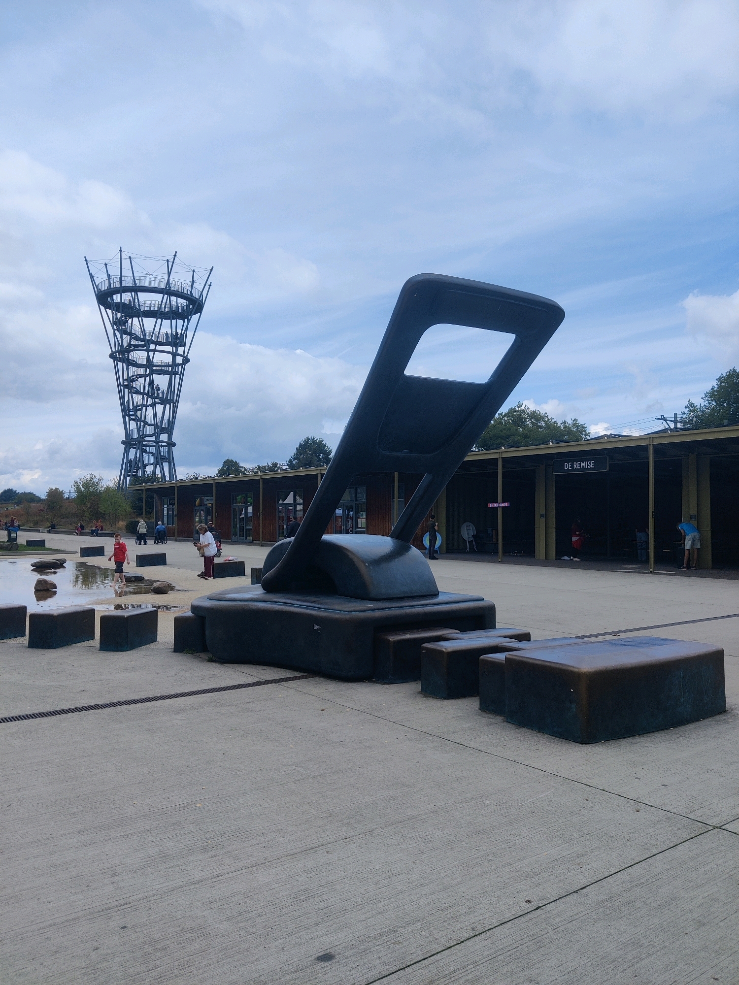 The image depicts an outdoor public space with a large, dark-colored abstract sculpture resembling a hole punch in the foreground. The sculpture is positioned on a concrete surface with rectangular block-like seating or decorative elements surrounding its base. In the mid-ground, a low-lying building with a covered walkway and signage that reads "DE REMISE" is visible. Several people are present in front of and within the building, some appearing to be patrons. To the left of the building, a tall, intricate metal observation tower with a spiraling staircase is prominent against a cloudy sky. In the foreground, to the left of the sculpture, there is a shallow water feature with rocks scattered around it, and a young boy in a red shirt is standing in the water. The overall setting suggests a park or recreational area. This is located at the **Houthavenpark in Tilburg, Netherlands**, which features the "De Pont" museum's sculpture garden and the new "De Pont" observation tower, often referred to as "De Ponttoren".