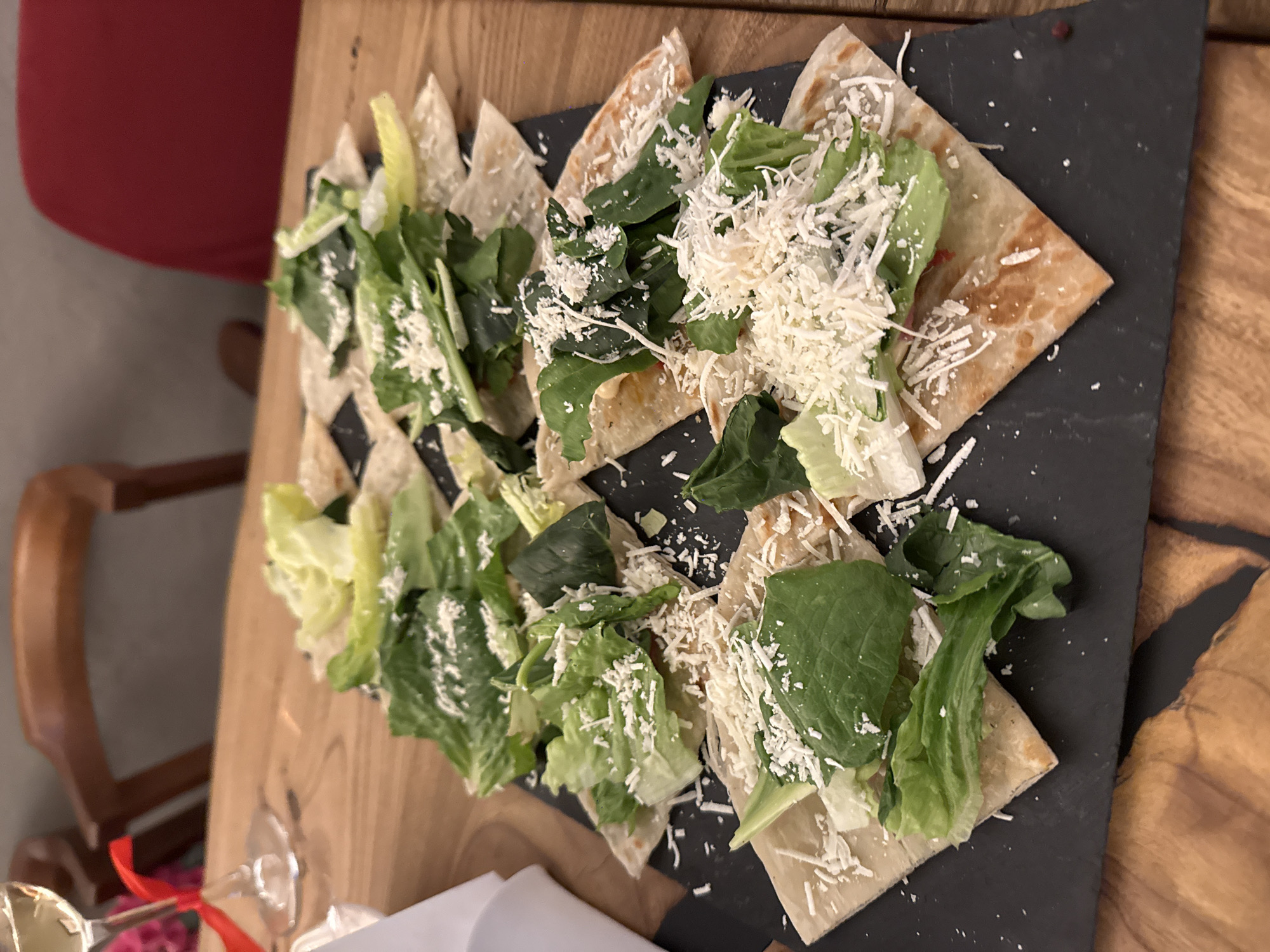 A close-up, slightly overhead shot captures a beautifully arranged dish on a wooden table, likely in a restaurant or cafe in Moscow, Russia. The main subject is a platter of food, presented on a rectangular black, slate-like serving board.
The dish consists of numerous triangular pieces of golden-brown flatbread or quesadilla, indicating they might be filled and lightly toasted. Each triangle is generously topped with a fresh salad mix, featuring both vibrant green Romaine lettuce and darker, possibly spinach or arugula leaves. A substantial amount of finely shredded white cheese, resembling Parmesan, is sprinkled over the greens, adding a contrasting texture and color. The flatbread pieces are artfully fanned out across the platter, creating an appealing visual spread.
The wooden table beneath the platter has a warm, medium-brown tone with visible grain, contributing to a cozy and inviting atmosphere. In the background, a blurred reddish object, possibly the back of a chair or an item of clothing, is visible on the upper left. Towards the lower left, parts of a wooden chair arm and a clear glass with a red ribbon tied around its stem suggest a celebratory or special dining occasion.
The overall scene depicts an indoor dining setting, emphasizing a focus on fresh ingredients and attractive food presentation. There is no visible text in the image.