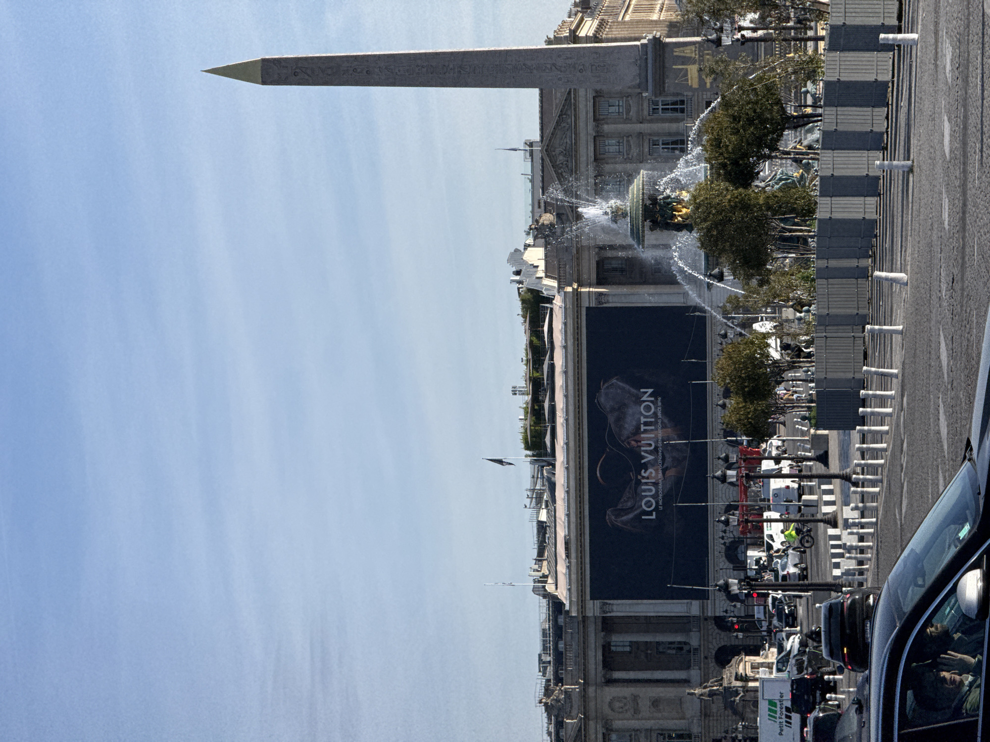 The image shows the Place de la Concorde in Paris, France. The Luxor Obelisk dominates the view. A fountain is visible, with water spraying. A large Louis Vuitton advertisement is displayed on a building. Traffic, including cars and a motorcycle, is visible. Pedestrians are present in the background. The sky is clear and blue.