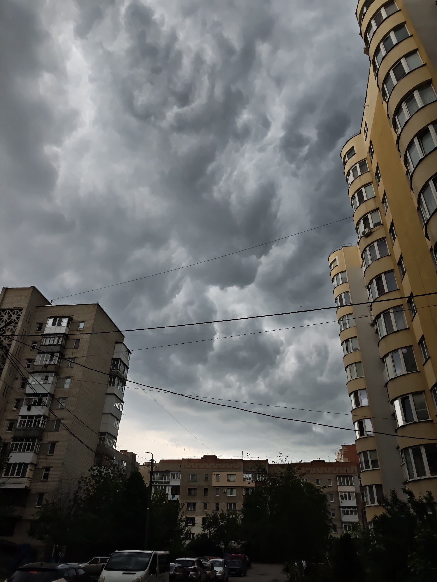 The image captures a street scene in Vinnytsia, Ukraine, under a stormy, overcast sky. Tall, multi-story apartment buildings flank the street. The building on the right is a curved, yellow apartment block, while the one on the left is a gray, rectangular building with exposed balconies and patterned concrete panels on one side. In the middle ground, lower apartment buildings with red-tiled roofs are partially obscured by trees. Power lines stretch across the sky, adding to the cluttered urban landscape. A row of parked cars and vans is visible at street level in the foreground. No people are visible in the frame. The atmosphere suggests an impending heavy rain or storm.