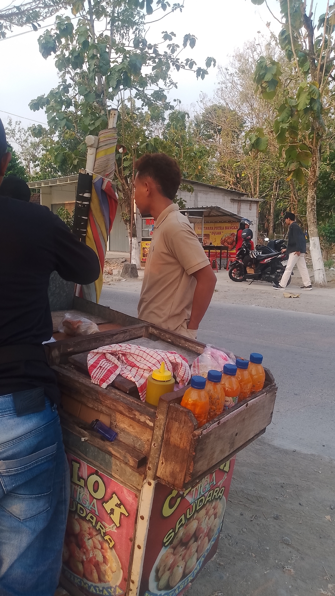A street food cart is displayed in the city of Karanglangu, Indonesia. The cart is made of wood and has a checkered cloth, a yellow squeeze bottle, and several orange bottles of sauce on top. The front of the cart features colorful signage with images of food and text that reads "LOK" and "TIGA SAUDARA". Two men are visible; one, wearing a black shirt and blue jeans, is partially obscured from view, and the other, in a tan polo shirt and khaki pants, stands facing away from the camera, looking towards the street. In the background, there is a road with a motorcycle and two more men, as well as several buildings and trees. The sky is overcast.