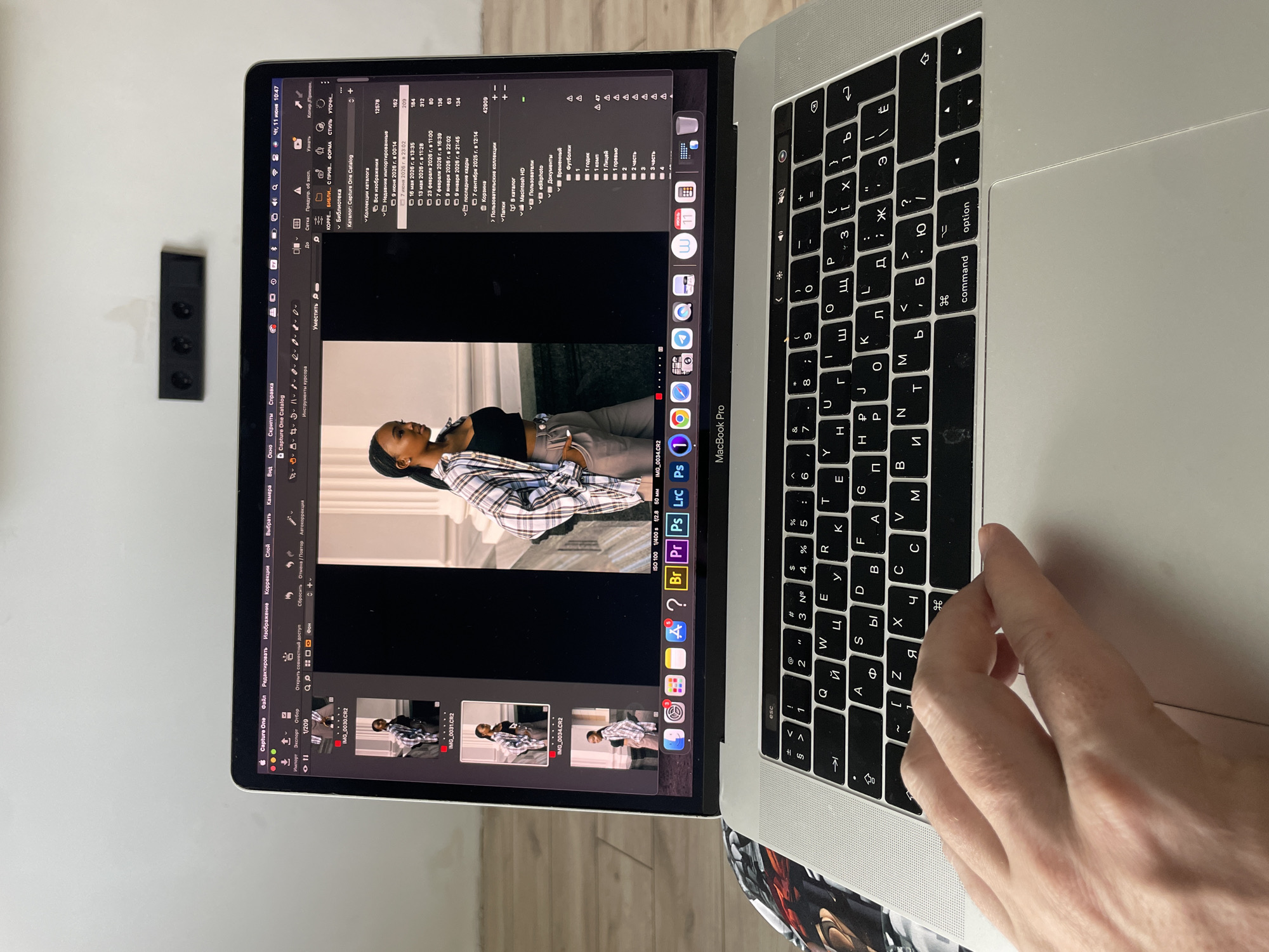 The image shows a close-up of a MacBook Pro laptop with its screen open. The laptop is displaying Capture One software, a photo editing application. On the left side of the screen, a filmstrip view shows several images of a young Black woman, with the currently selected image, "IMG_0034.CR2," prominently displayed in the center of the screen. The woman is wearing a plaid shirt over a black top and light-colored pants.
The right side of the screen shows catalog and library management tools within Capture One. The operating system language appears to be Russian, indicated by menu items like "Файл" (File), "Редактировать" (Edit), "Изображение" (Image), "Коррекция" (Correction), "Слой" (Layer), "Выбрать" (Select), and "Камера" (Camera). The date and time displayed are "Чт, 11 июня 10:47" (Thursday, June 11, 10:47).
A person's hand is resting on the laptop's trackpad. The background is blurred but appears to be an indoor setting with a light-colored wall and wooden flooring. There is also a black power strip mounted on the wall behind the laptop.
The prompt mentions the city of Kazan, Russia, but there is no visual information in the image that specifically places the scene in Kazan. The Russian language interface is the only clue that might suggest a connection to Russia.