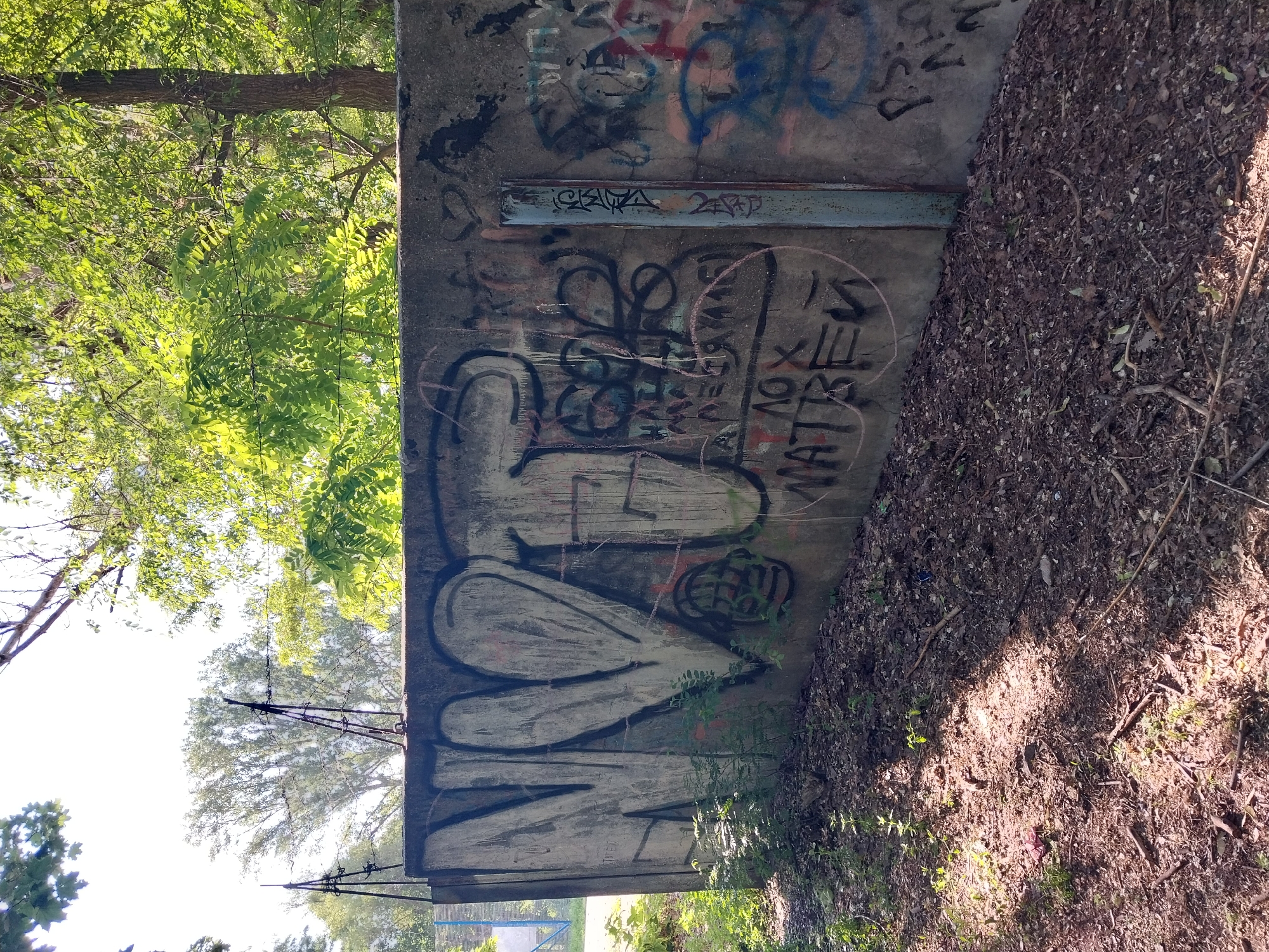 The image shows a concrete wall covered in graffiti, located in Dnipro, Ukraine. The wall is part of a fence topped with barbed wire. Green trees and foliage are visible behind the fence, with sunlight filtering through the leaves. The ground in front of the wall is covered in dirt and fallen leaves. The graffiti on the wall features large, stylized letters and symbols in various colors, including black, white, and red. Some of the graffiti appears to be spray-painted, while other markings seem to be done with markers or chalk. There are no people or animals visible in the image. The overall scene suggests an outdoor, urban environment with elements of decay and artistic expression.