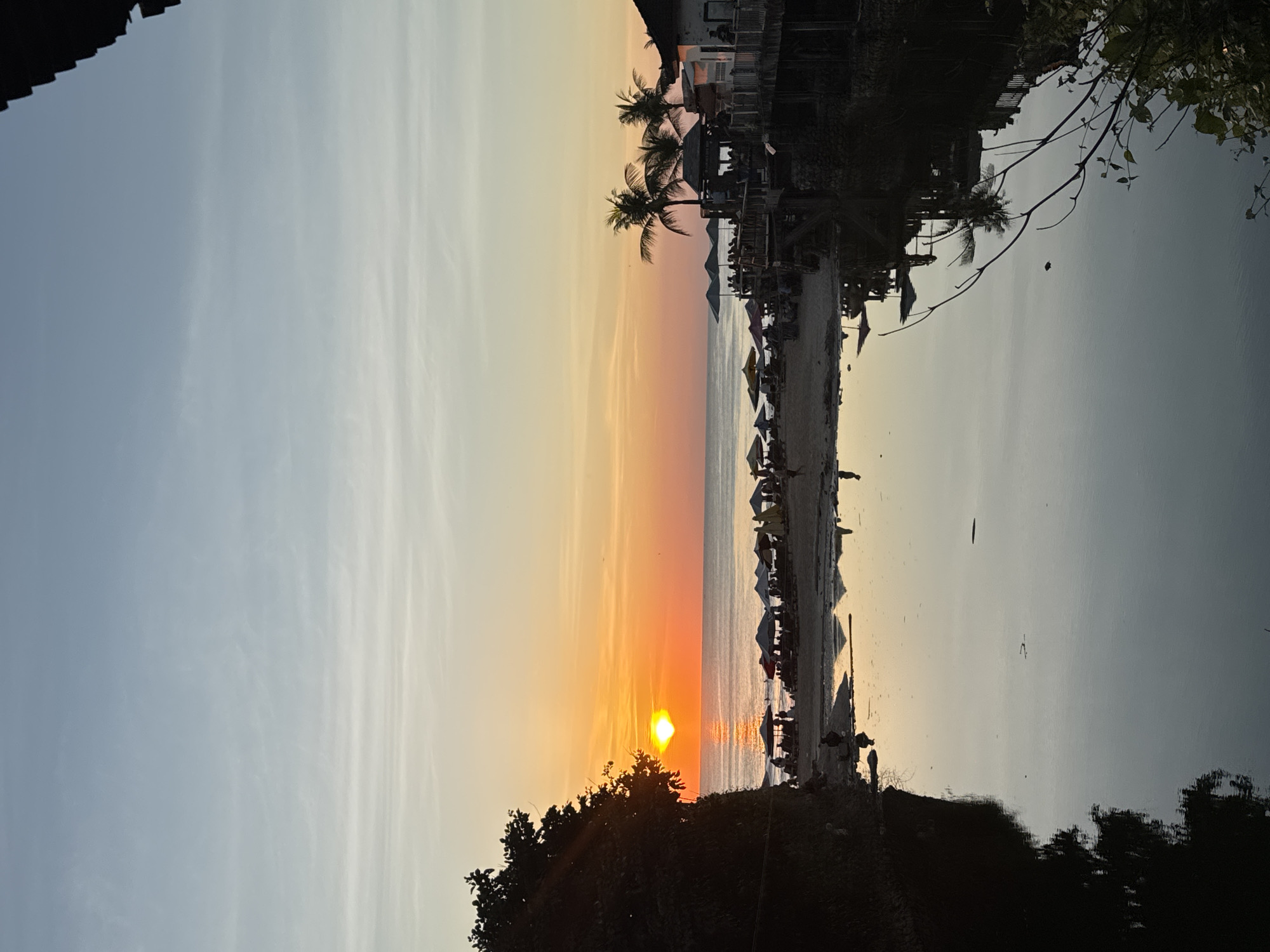 The image captures a sunset view over the ocean at a beach in Kuta Selatan, Indonesia. The sun is low on the horizon, casting an orange and yellow glow across the sky and reflecting on the water. Palm trees and beach umbrellas line the sandy shore. The silhouette of trees is visible in the foreground. There appear to be buildings and structures in the distance along the beach.