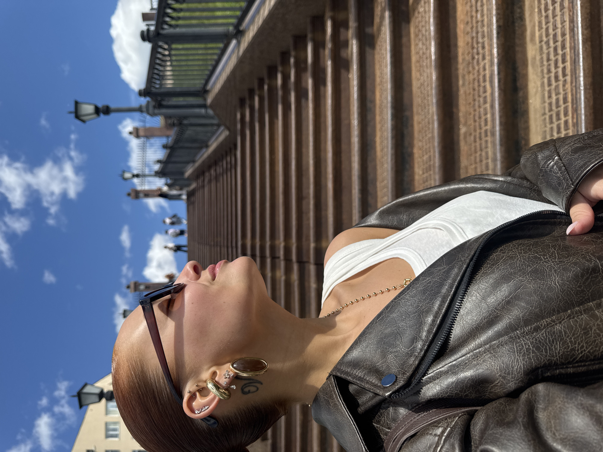 A young woman with auburn hair, a white tank top, and a dark brown leather jacket is posing in Izhevsk, Russia. She is on a set of steps with wooden slats along a metal railing near the Izhevsk State Circus. She wears dark sunglasses, gold hoop earrings, and has a tattoo near her ear. Behind her are passersby, buildings, and a blue sky with fluffy clouds.