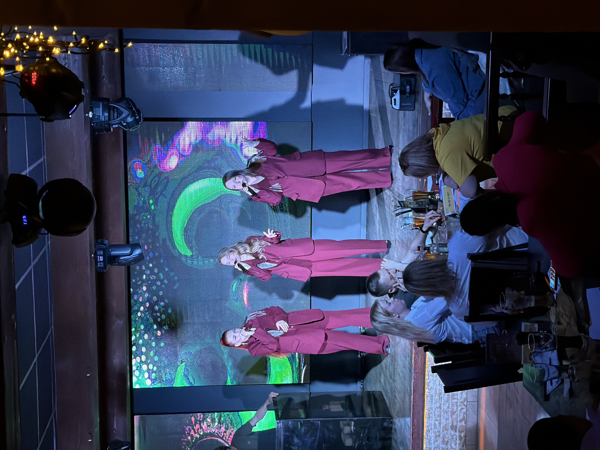 Captured in Chaikovsky, Russia, the image shows a dimly lit stage area with three women in matching red suits singing into microphones against a digital backdrop. The backdrop displays swirling colorful patterns. A lighting rig is visible above the stage. In the foreground, several people are seated at tables, observing the performance with drinks in front of them. Some are engaged in conversations. The setting suggests a live entertainment venue.