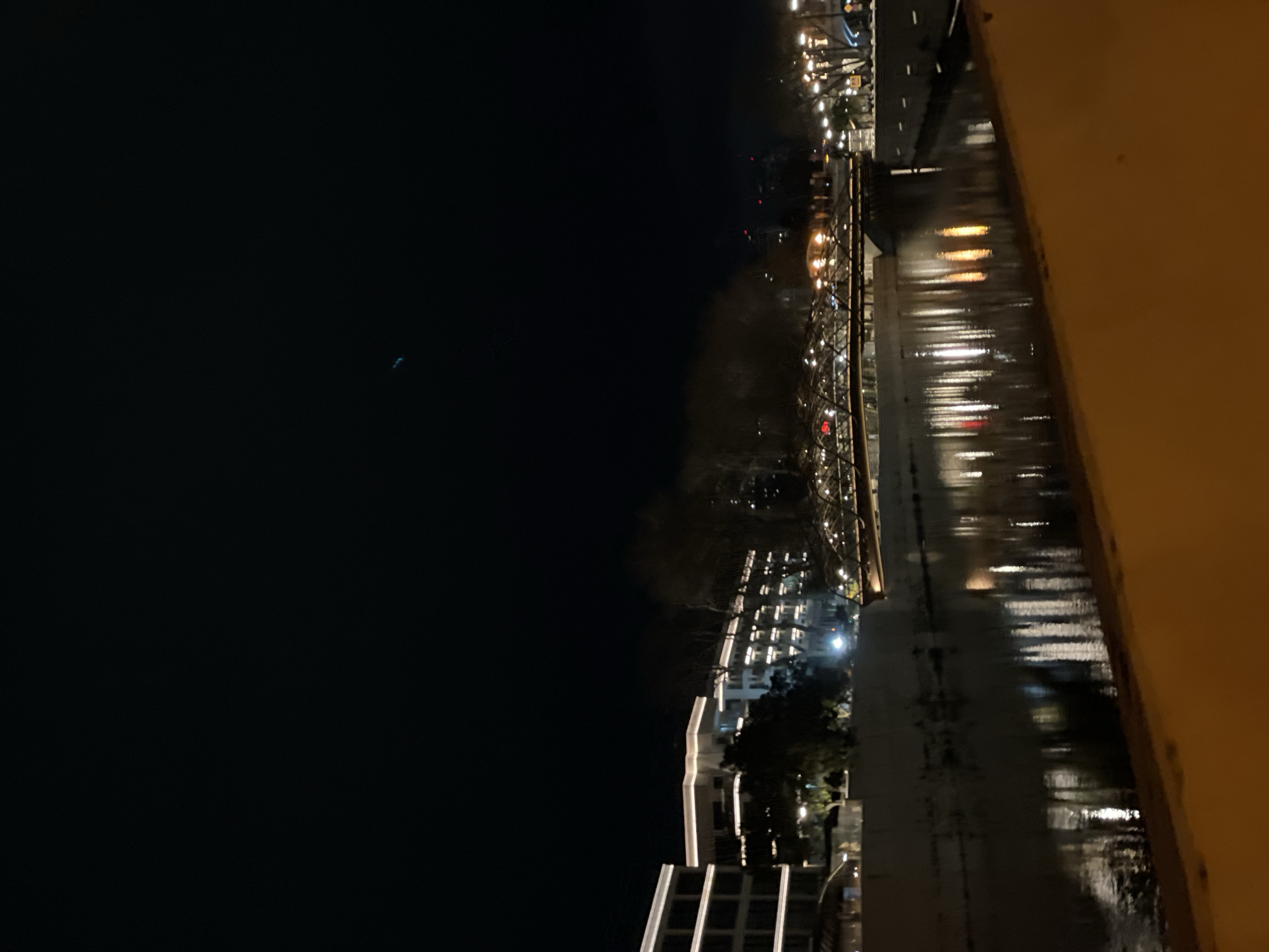 This image captures a nighttime urban waterscape in Sochi, Russia. A dark, calm body of water, consistent with a river or canal, flows through the center, reflecting numerous artificial lights from its surroundings.
On the left bank, multiple-story modern buildings are prominently lit with distinct horizontal and vertical white and yellow strip lighting along their facades. Bare-branched trees are visible between these structures and the water's edge.
A decorative bridge, possibly a pedestrian crossing, spans the waterway in the midground, heavily adorned with warm-toned string lights along its structure. Beyond the bridge on the far bank, additional streetlights and architectural lighting are visible. Two faint red lights appear in the distant background above the horizon line.
The foreground on the right consists of a broad, flat surface, likely a concrete embankment or walkway, which is strongly illuminated by an orange-yellow light source originating from outside the frame. The sky above is dark, with a faint, thin blue streak discernible in the upper-center portion. No human figures are present in the scene.
