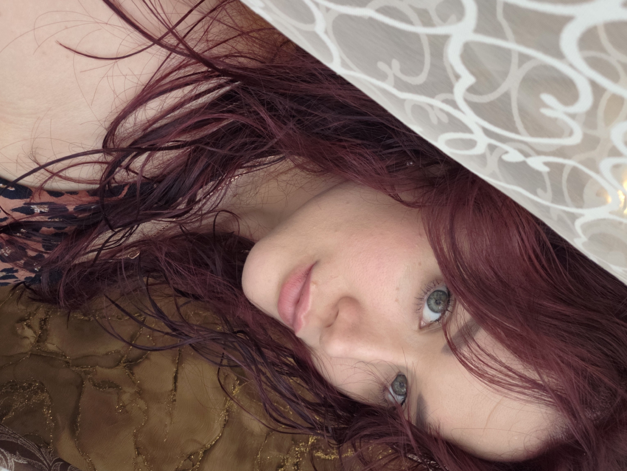 The image shows a close-up portrait of a woman with dark red hair and green eyes. She is partially covered by a sheer white fabric with a patterned design. Her head is tilted, and she is looking towards the camera. The background consists of a textured brown surface. There is no identifiable location within the city of Chaikovsky, Russia. The scene depicts a woman in a personal setting.