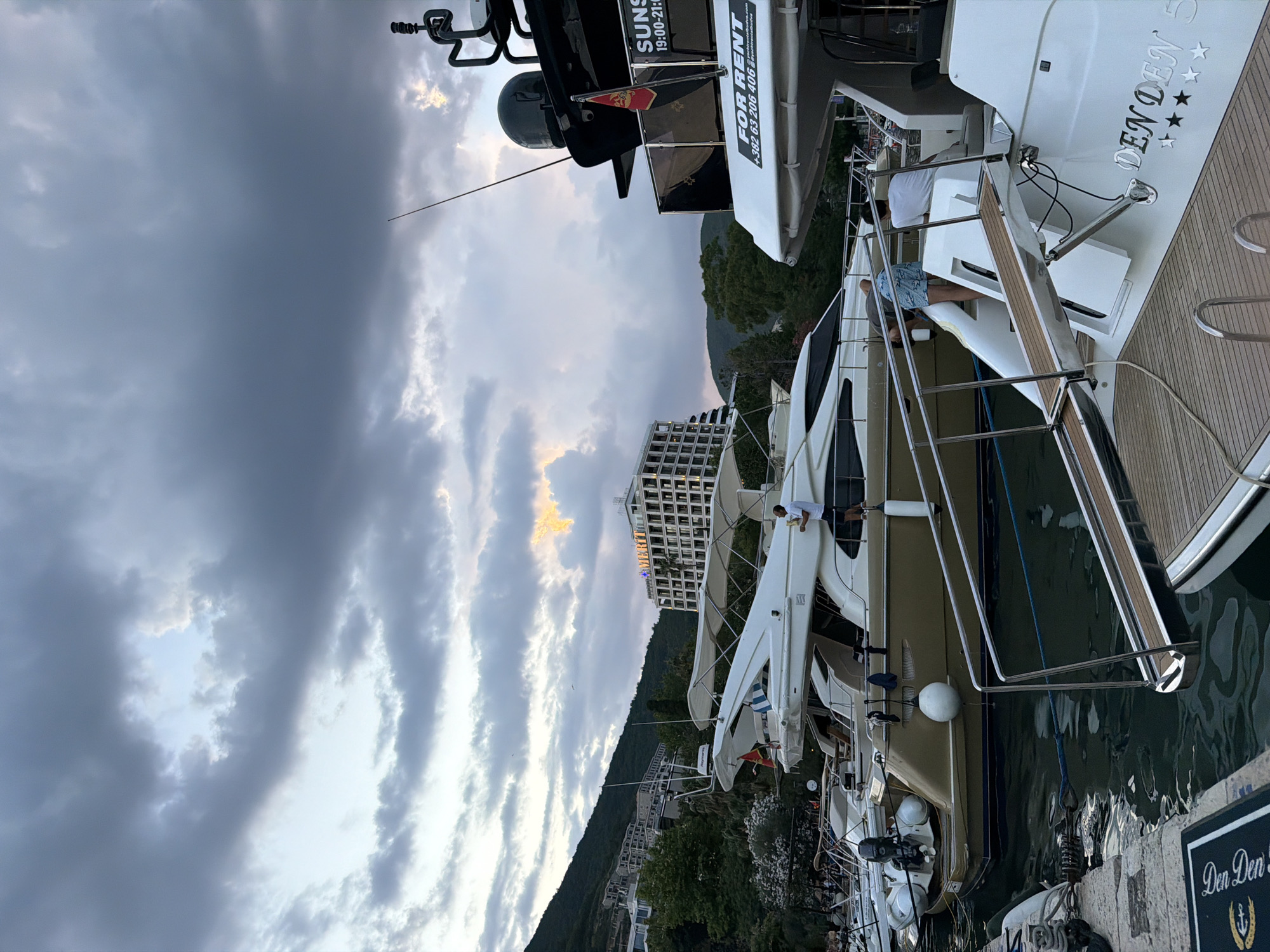 The image shows a marina with several luxury yachts docked. The sky is cloudy with hints of sunset colors peeking through. In the foreground, a large white yacht has "Den Den 5" written on its side in silver lettering. The yacht has a wooden deck and stainless steel railings. A person in blue shorts and a grey t-shirt is visible on the deck of this yacht, appearing to be relaxing. Another yacht, mostly black with white accents, is docked next to it, displaying text that reads "SUNS 19:00-21:00" and "FOR RENT +382 63 206 406". In the background, a large hotel with many windows is visible on a hillside covered in greenery, suggesting a resort or coastal town setting. The overall impression is a serene evening at a harbor, with the yachts implying leisure and potential for charter. The presence of a Montenegrin flag on one of the yachts subtly hints at the location being Montenegro.
