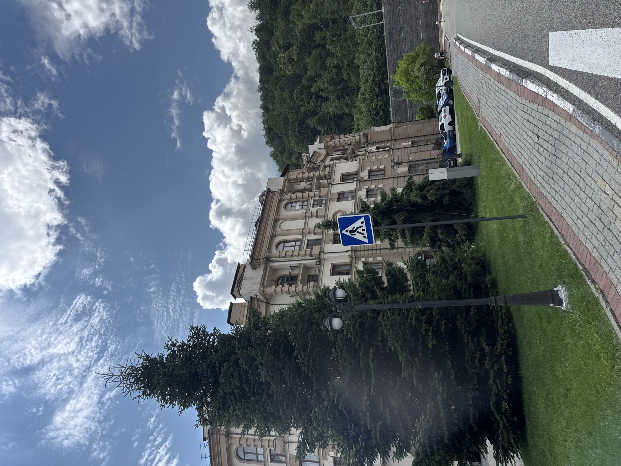 In Estosadok, Russia, a paved walkway with brick edging runs alongside a manicured lawn and a multi-story building. The building, with its beige facade and arched windows, appears to be a hotel or residential complex. A tall evergreen tree stands prominently in front of the building, partially obscuring it. A pedestrian crossing sign, a black lamppost, and several parked cars are also visible. The background is dominated by a dense, green forest on a hillside, under a bright blue sky with scattered white clouds.