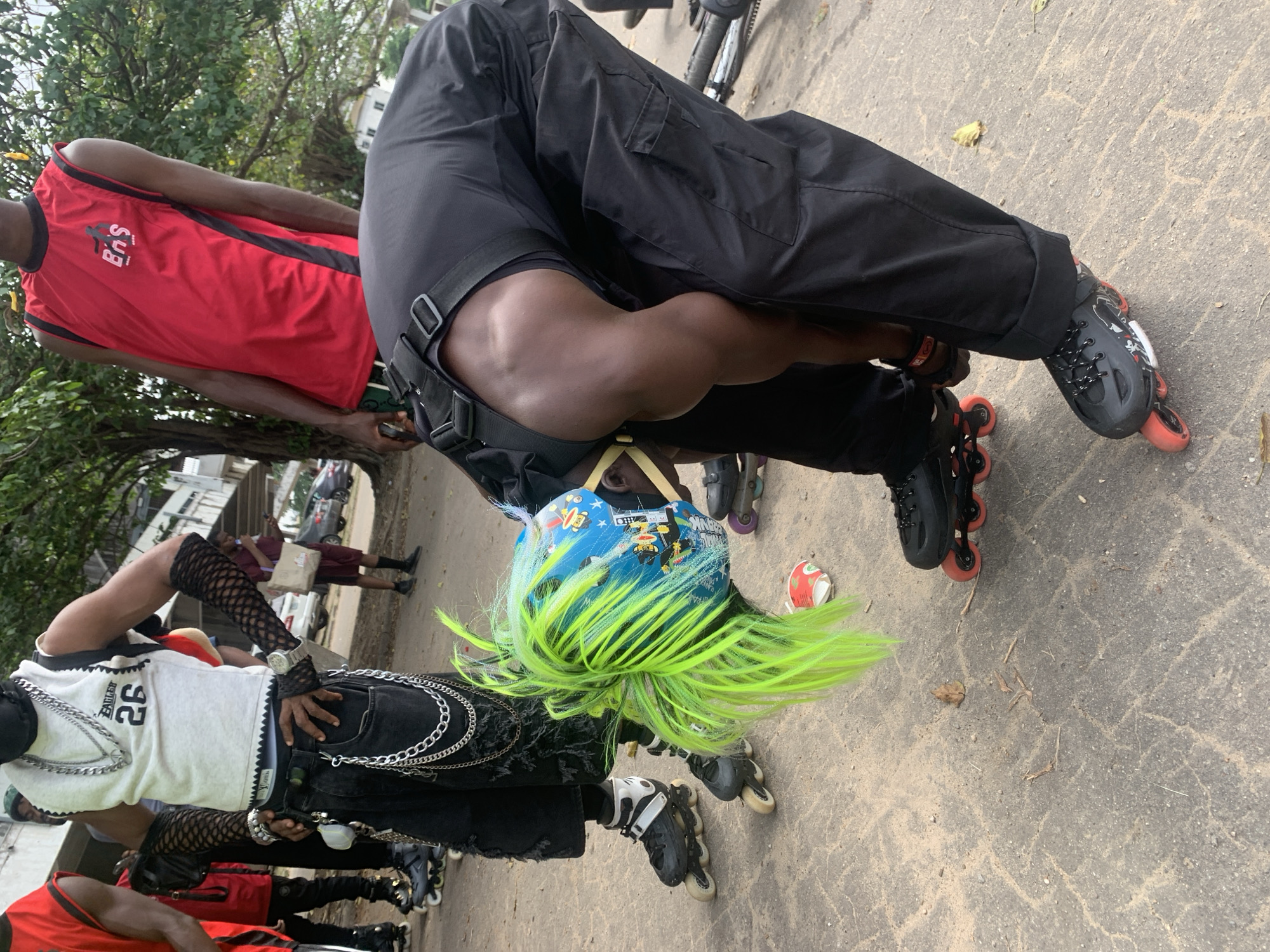 The image depicts an outdoor scene in Alagbede, Nigeria, likely a street or paved area adjacent to a building and trees. In the foreground, a person wearing black cargo pants and rollerblades is bending over to adjust their skates. This individual has bright green, spiky hair that extends from under a blue and yellow patterned helmet. They are also wearing a black vest with straps and a black face covering.
Behind this central figure, another person stands wearing a white sleeveless shirt with the number "26" on it, black ripped jeans, and black fishnet arm sleeves. They are adorned with multiple silver chains and bracelets. To the right, a third person is visible wearing a red sleeveless shirt with black trim and a logo that reads "BNS". Other individuals are partially visible in the background, some wearing rollerblades, and a person in a burgundy shirt and white bag is walking away. Several cars are parked in the background, suggesting an urban or semi-urban setting. The ground is a mix of cracked concrete and sand, with some dry leaves and debris scattered around. The overall atmosphere appears casual and possibly related to a gathering or sporting event involving rollerblading.