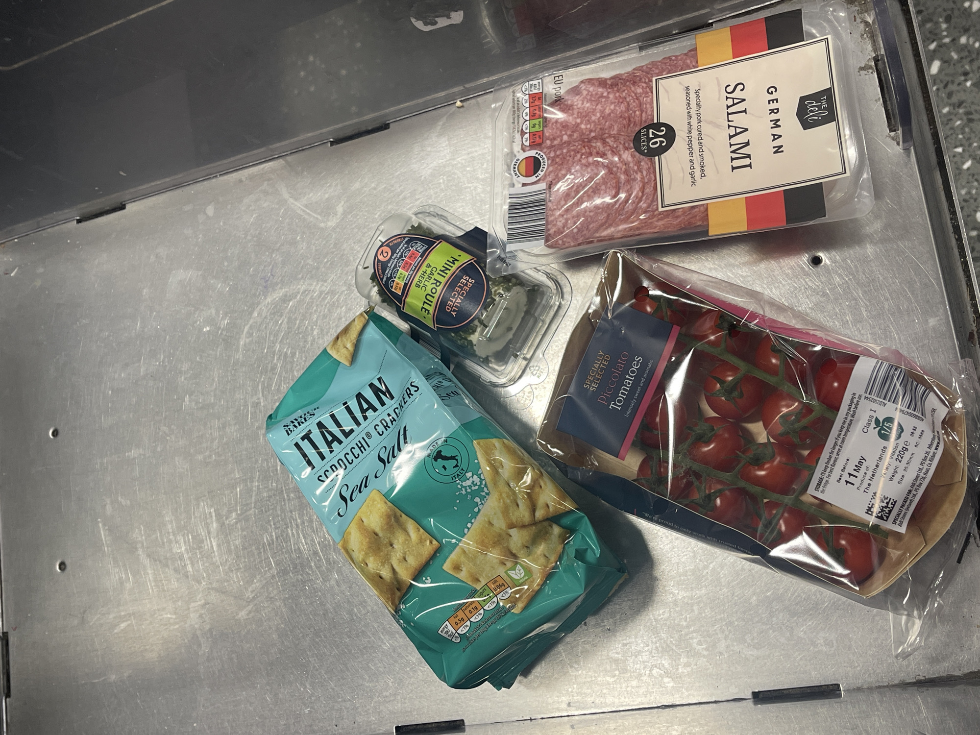 The scene is a close-up shot of a metal surface, likely a checkout conveyor belt, containing a selection of food items. These include a package of "Italian Scrocchi Crackers Sea Salt", a package of "Mini Roule Garlic & Herb" cheese, a package of "German Salami 26 slices", and a package of "Specially Selected Piccolata Tomatoes". The image contains no people and the location is not specific enough to determine which part of London the image was taken in.