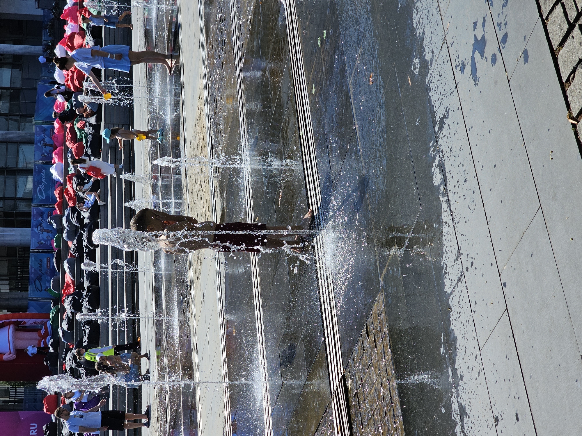 The image depicts an outdoor fountain area with several people, primarily children, interacting with the water jets on a sunny day. In the foreground, a young boy is seen partially obscured by water spray, wearing dark shorts. To his right, a young girl in a blue dress and yellow bucket is playing in the fountain, with an adult woman in a blue dress and white sneakers nearby, also holding a yellow bucket and seemingly supervising. Further back, on tiered steps, several people are seated on large beanbag chairs in various colors, including red, black, and white. Some individuals are standing and observing. The overall setting appears to be a public plaza or park, likely a modern urban space designed for recreation. The presence of beanbag chairs suggests a temporary setup, perhaps for an event or festival. The architecture in the background is somewhat obscured by trees and banners, but hints at a large, modern building. Considering the context of similar public spaces with large beanbag setups and fountains, this scene is likely located at Gorky Park in Moscow, Russia, known for its summer installations and events.
