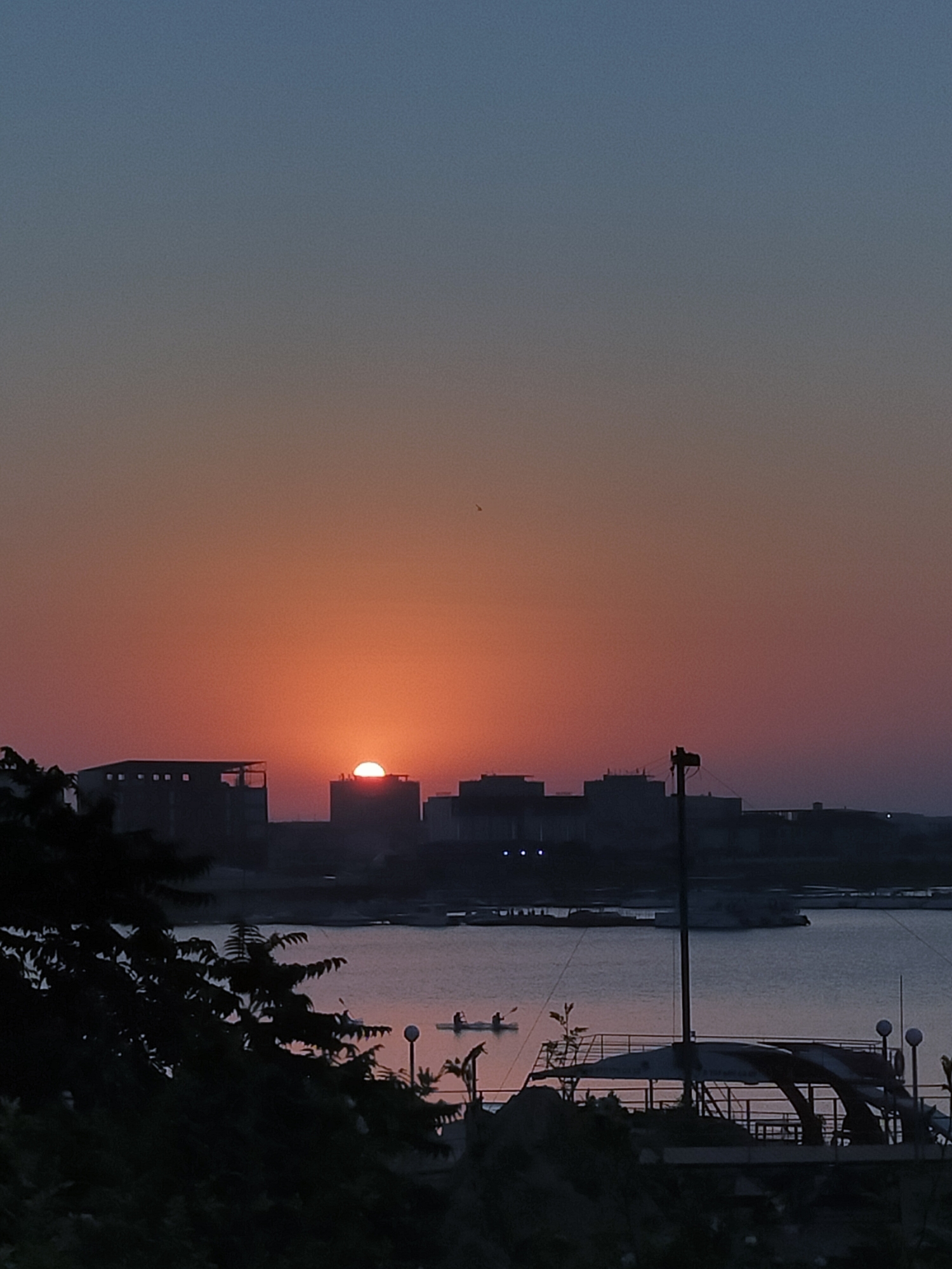 A sunset is depicted over the Caspian Sea in Aktau, Kazakhstan. The sun, a bright orange orb, is partially obscured by the rooftops of buildings in the foreground. The sky transitions from a soft orange at the horizon to a deep blue at the top. The water of the sea reflects the warm hues of the sunset. In the middle ground, several boats are visible on the water, with two individuals in one of them, appearing to be kayaking. Silhouettes of trees and a tower-like structure dominate the foreground, obscuring a portion of the view.