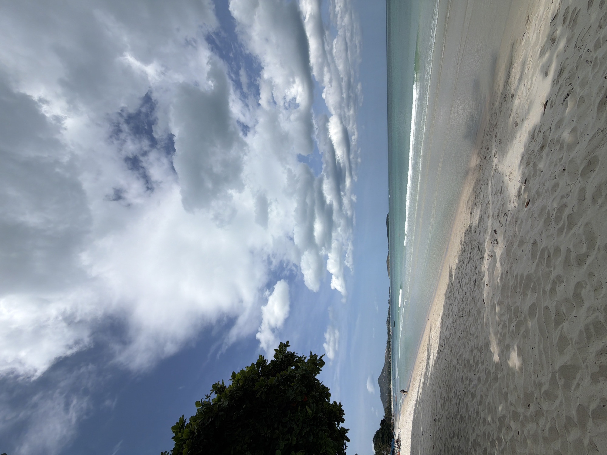 This is a photograph of a tropical beach scene, likely on the island of Ko Samui, Thailand. The image is dominated by a vast expanse of sandy beach in the foreground, with the gentle waves of the turquoise ocean lapping at the shore in the midground. The water appears clear and calm, with a few small waves breaking. In the distance, there are a few small islands or peninsulas visible on the horizon, suggesting the island's geography.
The sky above is a striking combination of bright blue and dramatic, billowing white and gray clouds. Some of the clouds are thin and wispy, while others are dense and puffy, casting shadows on the sand and water. The lighting suggests it is daytime, possibly late morning or early afternoon.
On the left side of the image, a large, leafy green tree provides shade, its branches reaching out towards the beach. Further down the beach, in the far distance, a few beach chairs and umbrellas can be seen, indicating a sparsely populated area. A couple of individuals are walking along the shoreline, their figures small against the vastness of the landscape.
The sand itself is light-colored and appears to have some texture, with small ripples and footprints indicating recent activity. Shadows from the tree and possibly other unseen objects are cast across the sand, adding depth and dimension to the scene. The overall atmosphere is one of tranquility and natural beauty, characteristic of a tropical paradise.