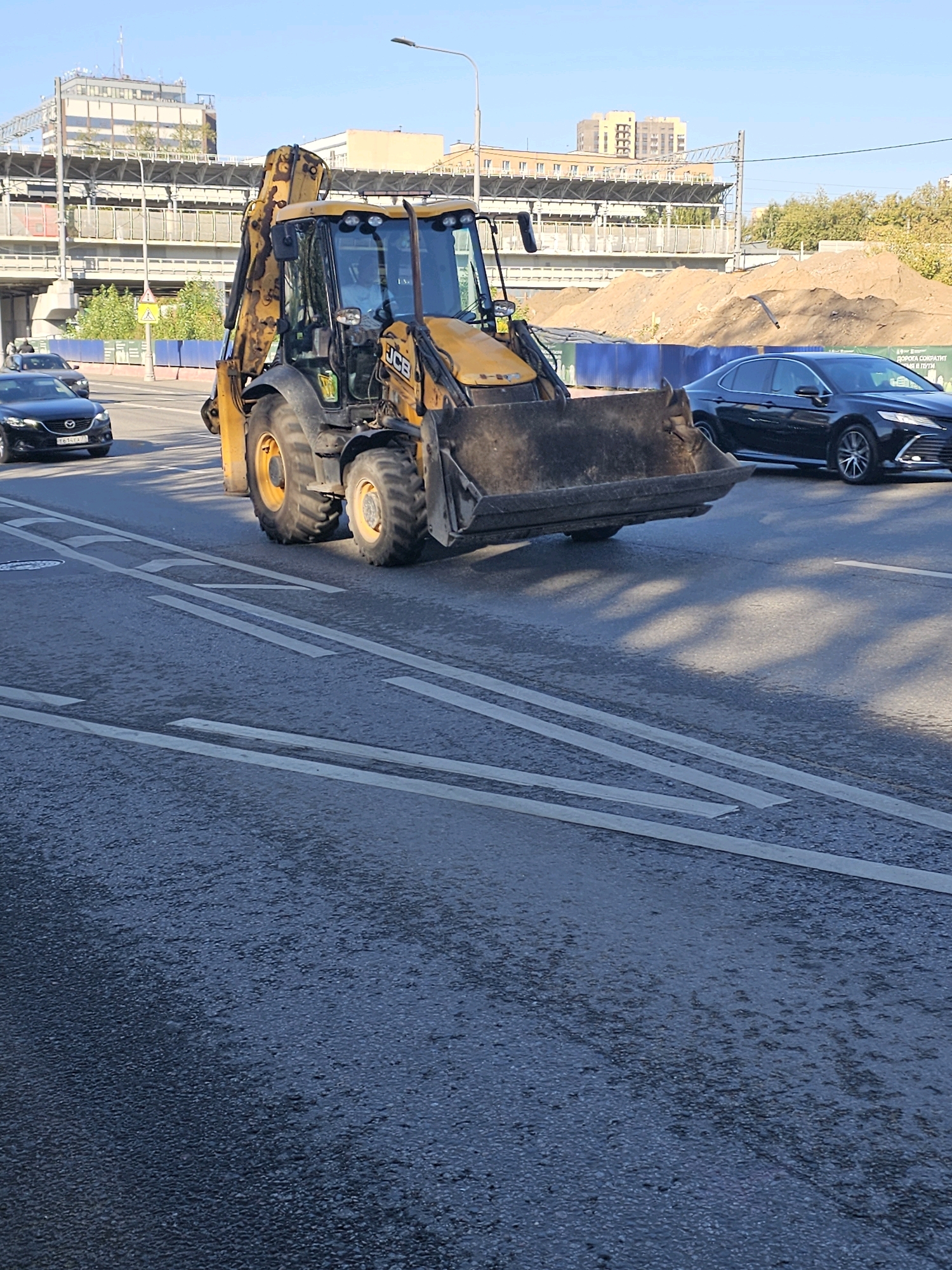 A yellow JCB backhoe loader is parked on an asphalt road in Moscow, Russia. The backhoe is positioned in the foreground, with its large bucket facing the viewer. The machine appears to be stationary. In the background, there is a black sedan in the adjacent lane and a construction site with piles of dirt and a blue fence. Elevated structures, possibly part of a railway or highway, are visible further in the distance, along with buildings and trees. The sky is clear and blue. The road surface shows visible lane markings. The scene appears to be captured during daylight, with shadows cast by the backhoe and foliage on the road.