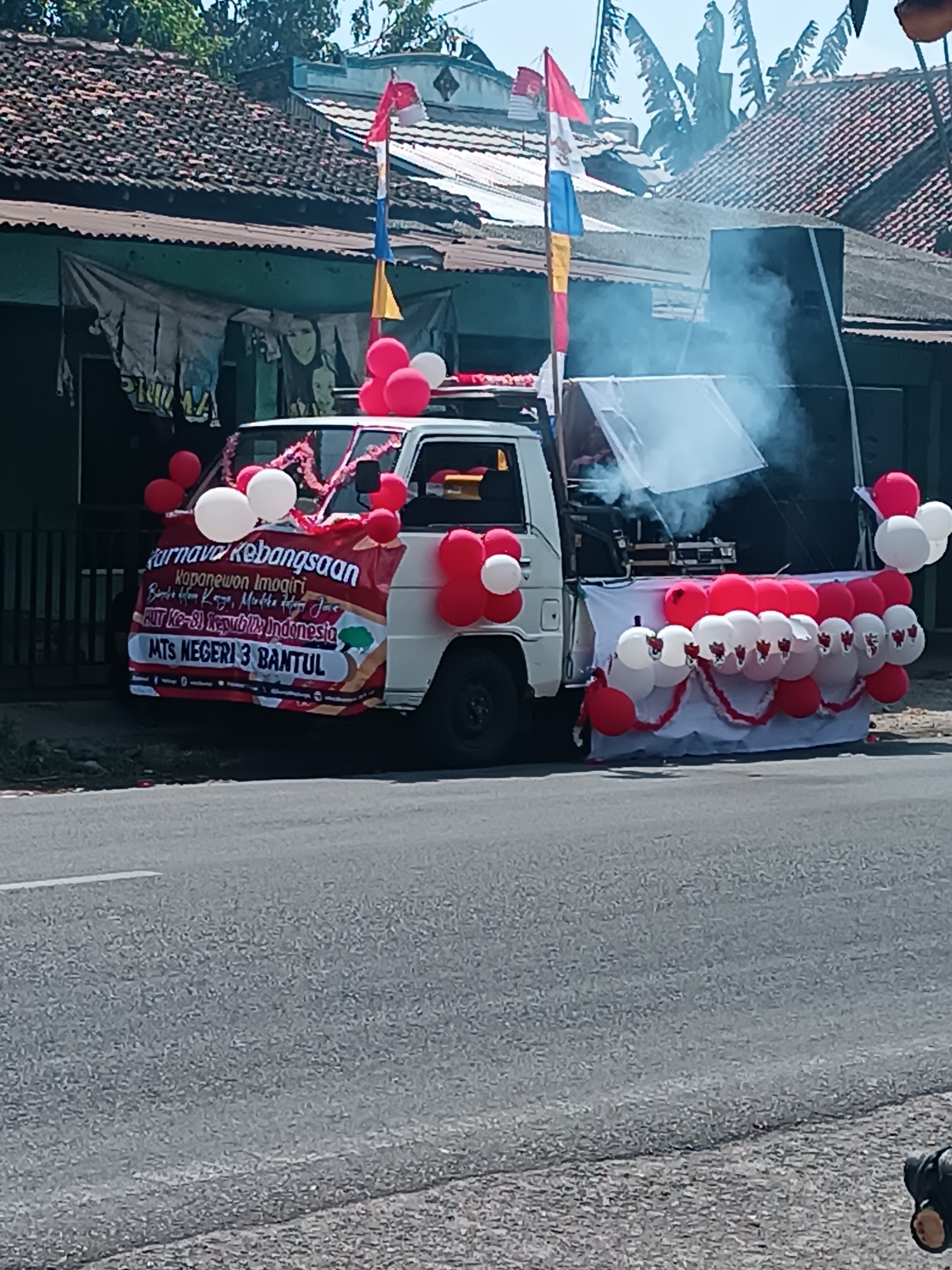 A white pickup truck is decorated with red and white balloons and banners for a "Carnaval Kebangsaan" (National Carnival) in Imogiri, Indonesia. The truck has a sound system on the back, with smoke coming from it. The banner reads "Kapanewon Imogiri, Bersatu Kita Kuat, Merdeka Hapy Jas HUT KE-69 Republik Indonesia MTS NEGERI 3 BANTUL". The truck is parked on a paved road in front of a building with a tiled roof, likely in the city of Imogiri. The sky is blue and sunny.