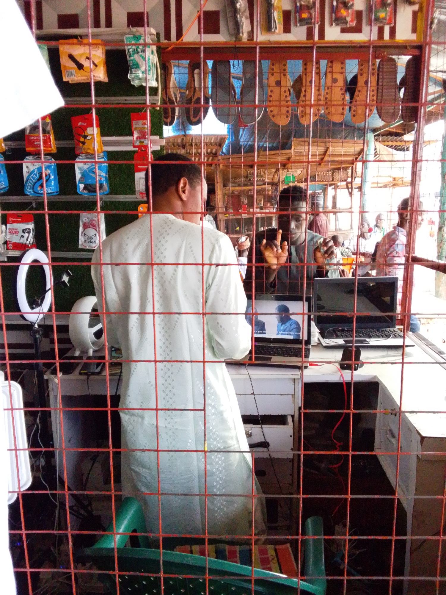 In Serti, Nigeria, a man wearing a light-colored, patterned garment stands facing away from the camera, his back to a metal security grate. He is positioned in what appears to be a market stall or small shop, with various items displayed behind him, including packaged electronics like chargers and headphones, as well as several pairs of sandals hanging from a rack. To his left, a ring light on a stand and a small white fan are visible on a counter. In front of him, two laptops are open on a desk. One laptop displays a video of two men. A second man, wearing a dark shirt, is visible through the grate, looking at the camera and holding up a smartphone. He appears to be interacting with the person operating the camera. Other people are visible in the background of the stall, some of whom are looking towards the front. The overall environment suggests a busy commercial setting within the city.