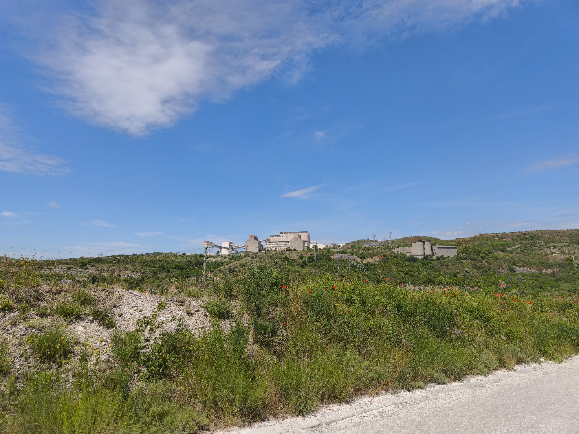 A landscape featuring an industrial complex in the distance under a blue sky with scattered clouds. The foreground consists of vegetation covering a rocky terrain, adjacent to a light-colored road. The industrial complex is situated on a hill, with various buildings and structures visible. The location is Unknown, Unknown.