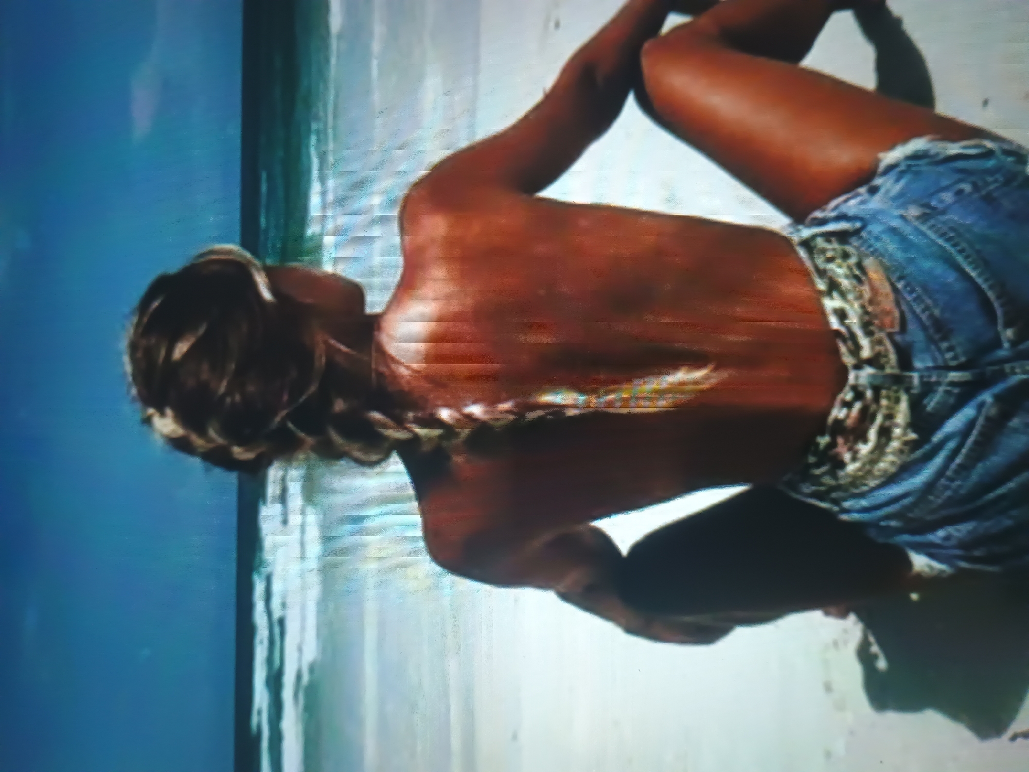 A person is depicted from behind, seated on a sandy beach, gazing towards the ocean under a clear blue sky. The individual exhibits deeply tanned skin with a reddish-brown hue across their back and shoulders, which are bare. Their hair is light brown and styled into a single, long braid extending down the spine, with lighter blonde strands visible towards the end of the braid. The person is wearing light blue denim cutoff shorts with frayed edges, secured by a patterned belt, which appears to be animal print, visible above the waistband. Their legs are bent, with the right knee elevated. In the background, a blue-green ocean stretches to the horizon, featuring several distinct lines of white-capped waves breaking towards the shore. The sky above is a consistent bright blue with minimal cloud cover. The location is a coastal beach environment in the city of Unknown, Unknown.