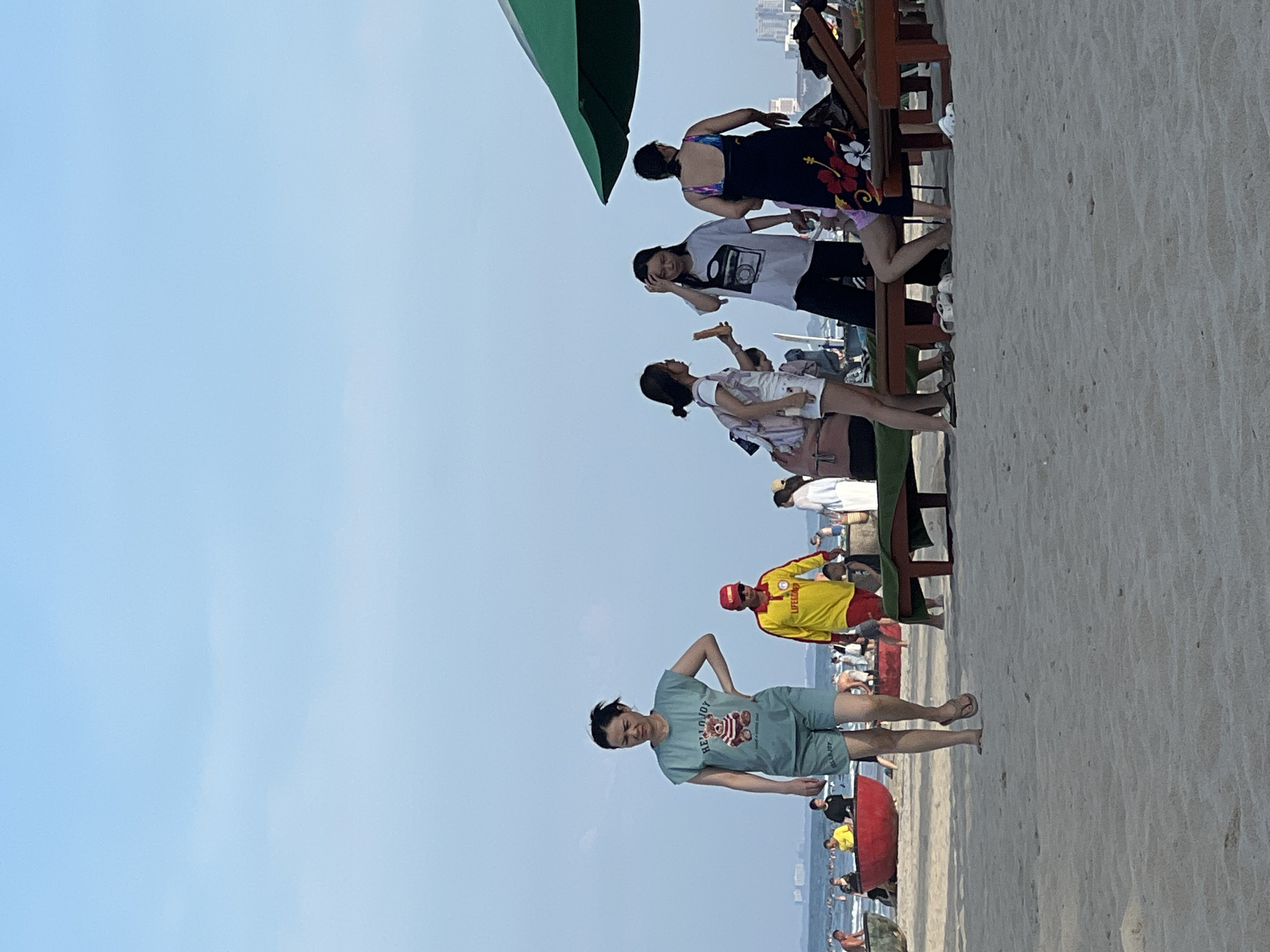 A group of people are gathered on a sandy beach in Da Nang, Vietnam, under a bright blue sky. In the foreground, a woman wearing a teal t-shirt with a teddy bear graphic and matching shorts walks towards the camera, her left hand on her hip. To her right, a man in a yellow and red lifeguard uniform with "LIFEGUARD" visible on his sleeve is walking away from the camera. Further in the background, several other individuals are visible, some sitting on lounge chairs under a large green umbrella, others standing or walking near the water's edge. Red fishing boats are positioned on the shore. The scene suggests a recreational day at the beach, with the presence of a lifeguard indicating a public swimming area.