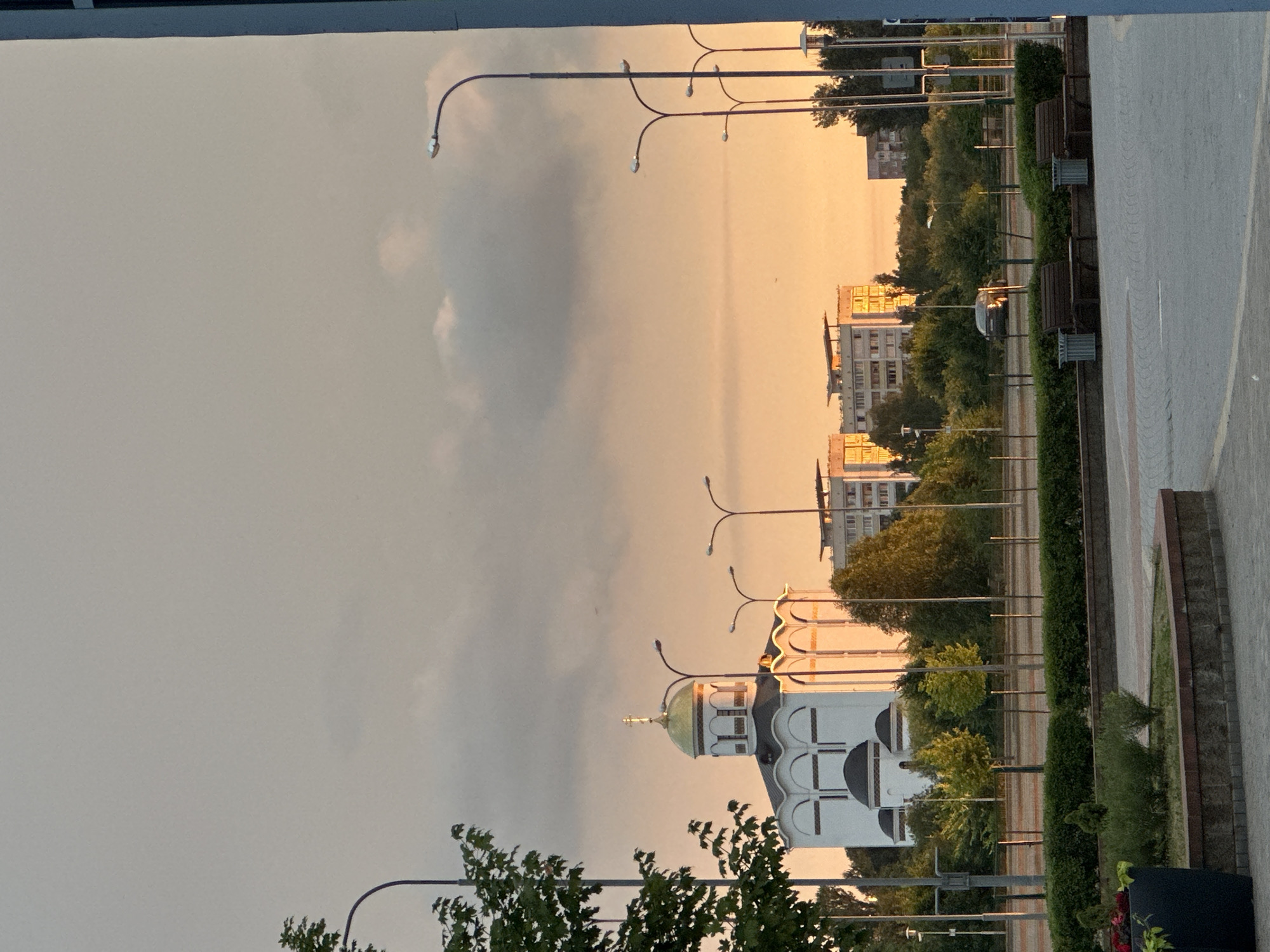 The image captures a view of the Church of All Saints in Minsk, Belarus, during a sunset. The sky is a gradient of gray and orange hues, with some scattered clouds. The church, a white building with a golden dome, stands prominently in the left half of the frame. It features multiple arched windows and a cross atop its dome. Several streetlights are visible in the foreground and midground.
Behind the church, modern apartment buildings with flat roofs are situated amidst lush green trees. The lower part of the image shows a paved area with planters, benches, and trash receptacles, suggesting a public space or park. A dark-colored car is parked on a road in the mid-ground. The overall scene conveys a peaceful urban landscape at dusk.