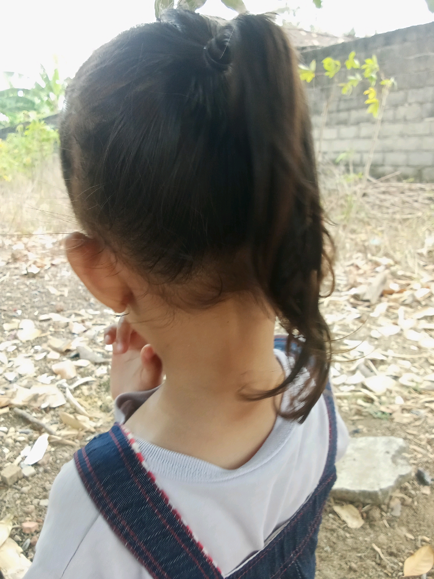 An over-the-shoulder, close-up shot shows the back of a young child's head and neck from a side perspective. The child's dark hair is pulled back into a high ponytail. The child is wearing a white shirt with dark blue overalls that have red trim. The child's right ear is visible, and their right hand is reaching up towards their face. The background is blurred, showing dry leaves and dirt on the ground, a brick wall, and some foliage. The scene appears to be outdoors, possibly in a backyard or a rural area of Unknown, Indonesia.