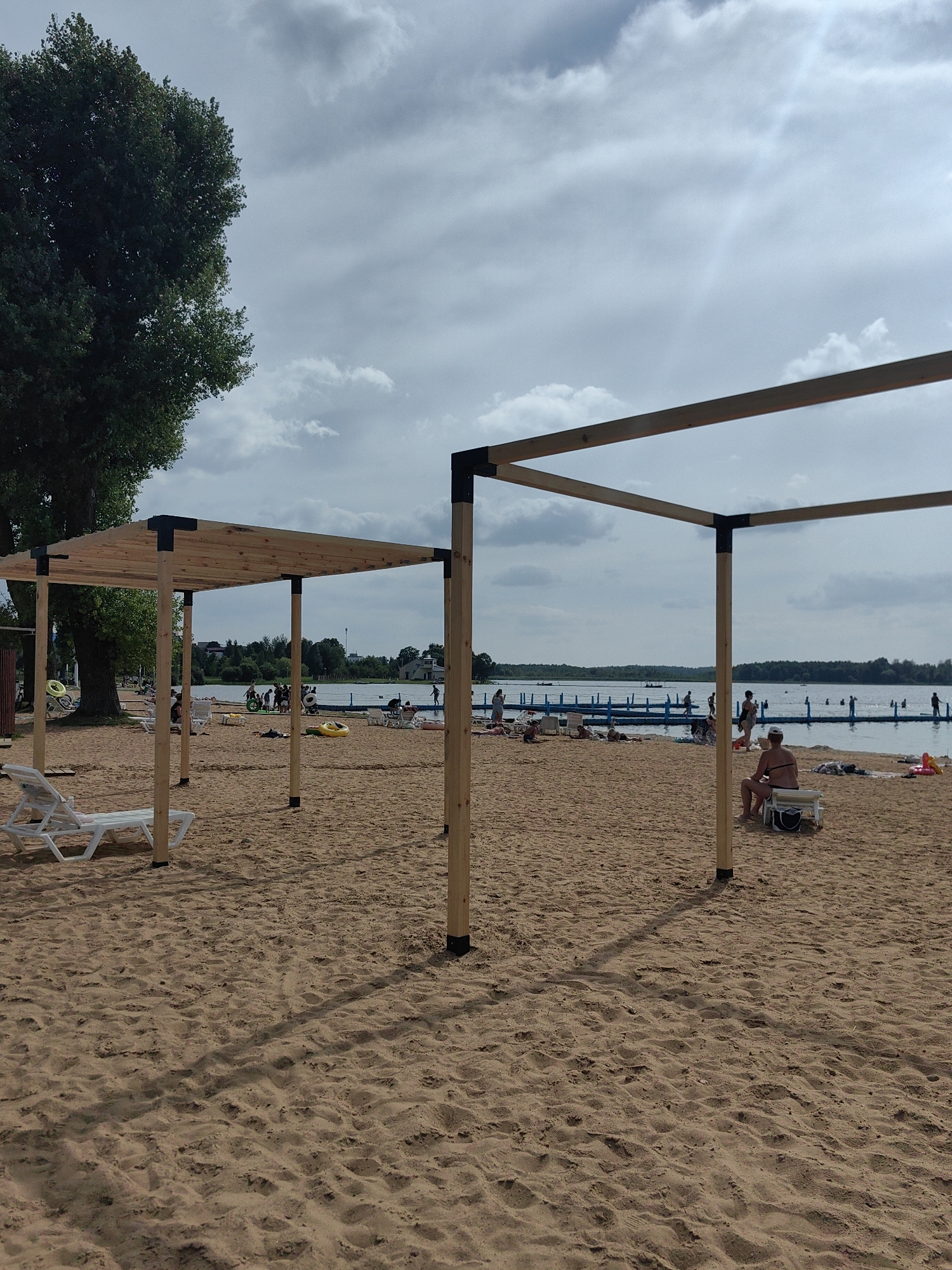 The image depicts a sandy beach area on a cloudy day, likely at Lake Myastro in Myadzyel, Belarus. Two unfinished wooden pergolas with dark corner connectors are prominent in the foreground, casting shadows on the sand. In the background, a body of water, presumably the lake, is visible with a floating platform structure and several people scattered around the beach and in the water.
On the left side of the image, a large, leafy tree stands tall. In the middle ground, there are lounge chairs, beach umbrellas, and inflatable toys. A person is seated on a lounge chair under the right pergola. Other individuals are seen walking, sitting, and swimming in the lake. The overall scene suggests a public recreational area during a summer day, with some infrastructure still under construction.