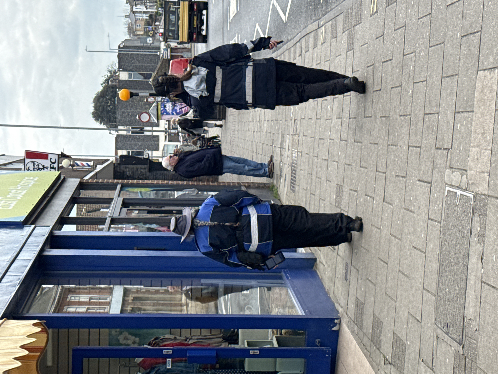 The image captures a street scene in Gorleston-on-Sea, United Kingdom. Two individuals are walking on a paved sidewalk alongside a blue-painted storefront with a display window. One individual is wearing a dark-colored jacket with white reflective stripes and dark pants, while the other is wearing a similar jacket but with blue accents. Both appear to be holding or using mobile phones. Further down the sidewalk, an older man in a dark jacket and jeans is walking in the same direction. In the background, there is a KFC sign, a barber pole, and various street signs and buildings, including a yellow bus or truck further down the road. The sky is overcast.