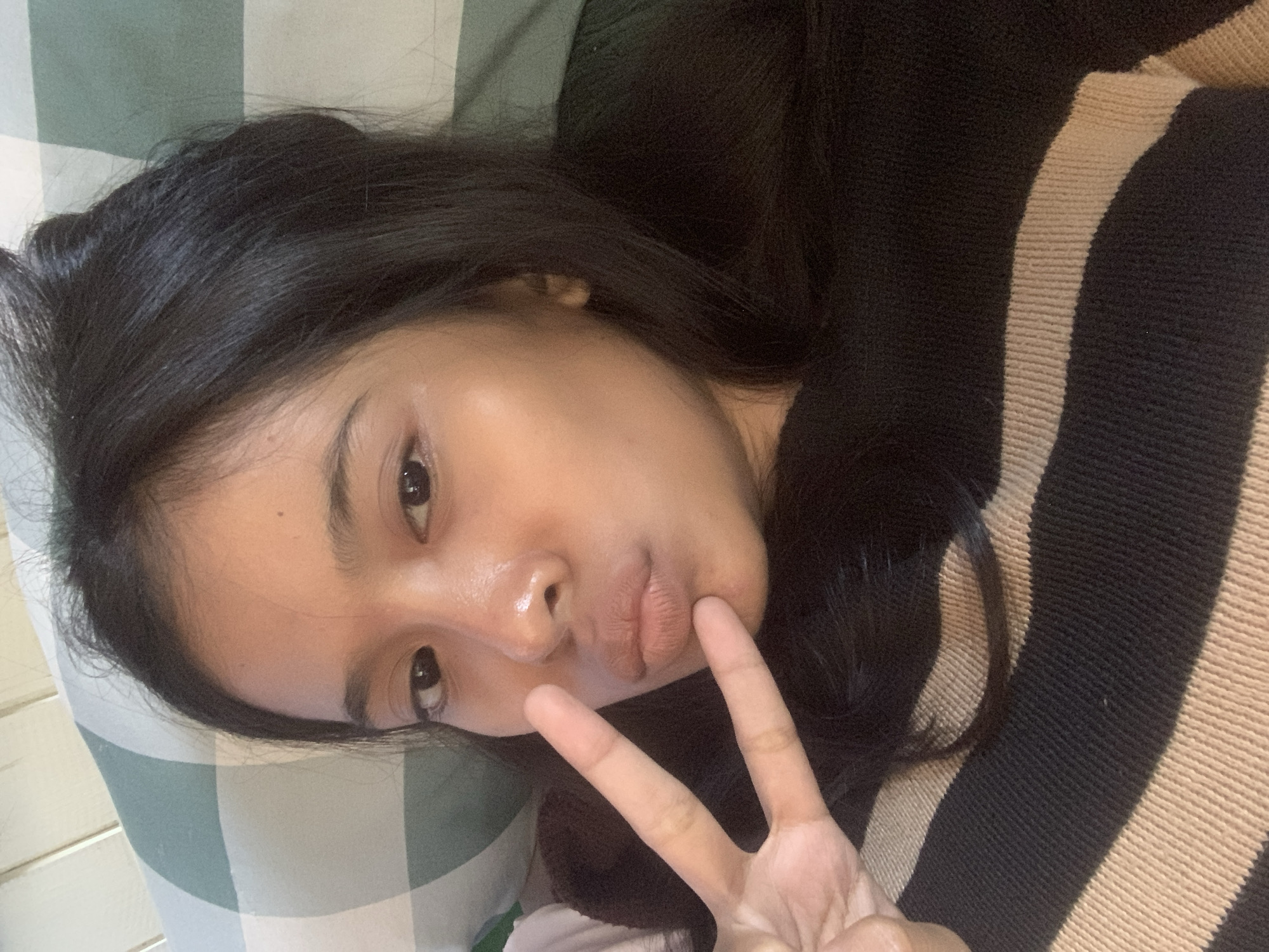 A young woman is captured in a close-up, horizontal selfie, lying down indoors in Sungai Meriam, Indonesia. She has long, dark hair cascading around her head and looks directly at the camera with a relaxed expression, her lips slightly pursed. Her right hand is raised, forming a peace sign. She is dressed in a black top or sweater featuring prominent horizontal beige stripes. The background behind her head reveals a green and white plaid-patterned pillow or blanket, suggesting she is on a bed or sofa. To her left, a light-colored wooden or paneled surface is partially visible. Her skin has a noticeable sheen, and the lighting is soft and ambient, consistent with an indoor setting. The overall mood conveyed is casual and friendly. No text is visible in the image.