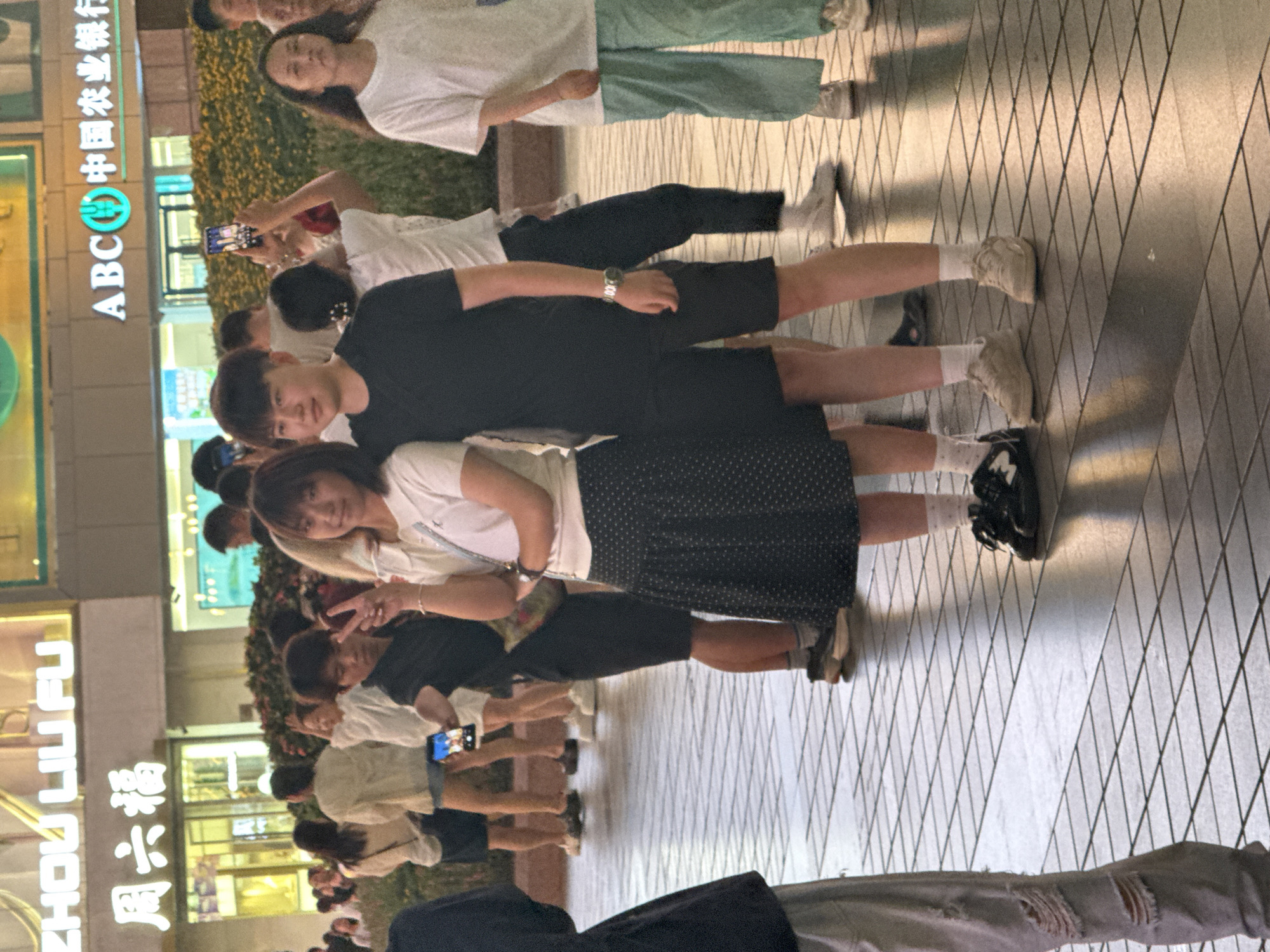 In the Yuzhong District of China, a group of young adults are gathered on a tiled pedestrian walkway, illuminated by artificial lights and signage from the surrounding commercial buildings. In the foreground, a young woman wearing a white polo shirt and a polka-dot skirt stands with her right hand raised, forming a peace sign, while a young man in a black t-shirt and shorts stands beside her. Both are looking towards the camera. Behind them, other individuals are visible, some holding up mobile phones, possibly to take pictures or record. The background features illuminated signs, including "ZHOU LIU FU," "周六福," and "ABC 中国农业银行," indicating a commercial area. The pavement is composed of square tiles laid in a diamond pattern, and shadows are cast by the people and the overhead lighting.