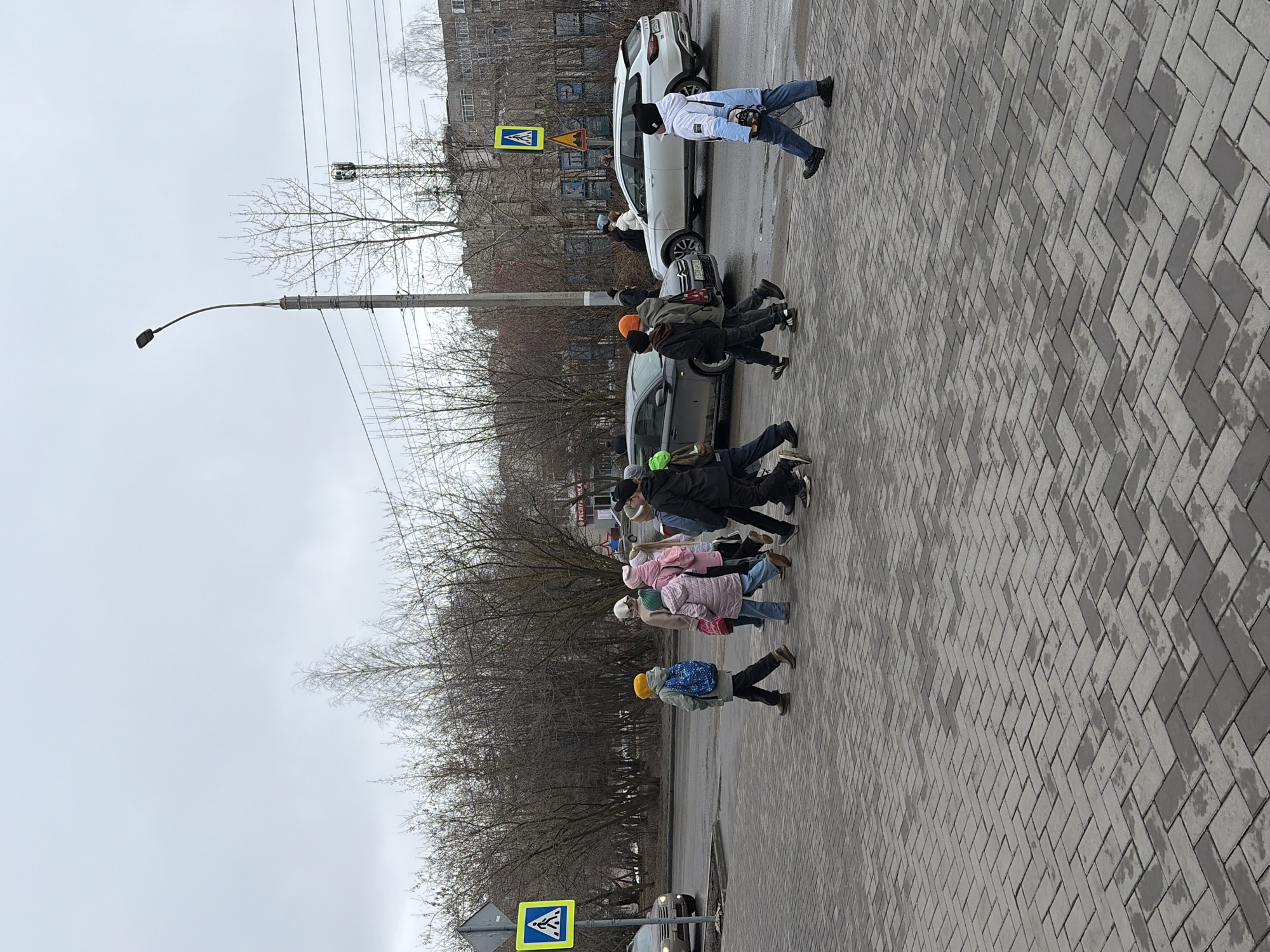 A group of people are crossing a pedestrian crosswalk on a paved road in what appears to be an urban environment. The location is Izhevsk, Russia.
There are several pedestrians, including children wearing brightly colored hats (yellow, green, orange, blue). They are proceeding across the crosswalk. Several adults are also present in the group.
A number of vehicles are visible, including a white SUV, a gray car, and other cars slightly visible further down the road. There are pedestrian crossing signs located on either side of the crosswalk.
The weather appears overcast or rainy. The trees along the roadside are bare, suggesting it is either late autumn or early spring. A lamppost stands near the crosswalk. Buildings are visible in the background.