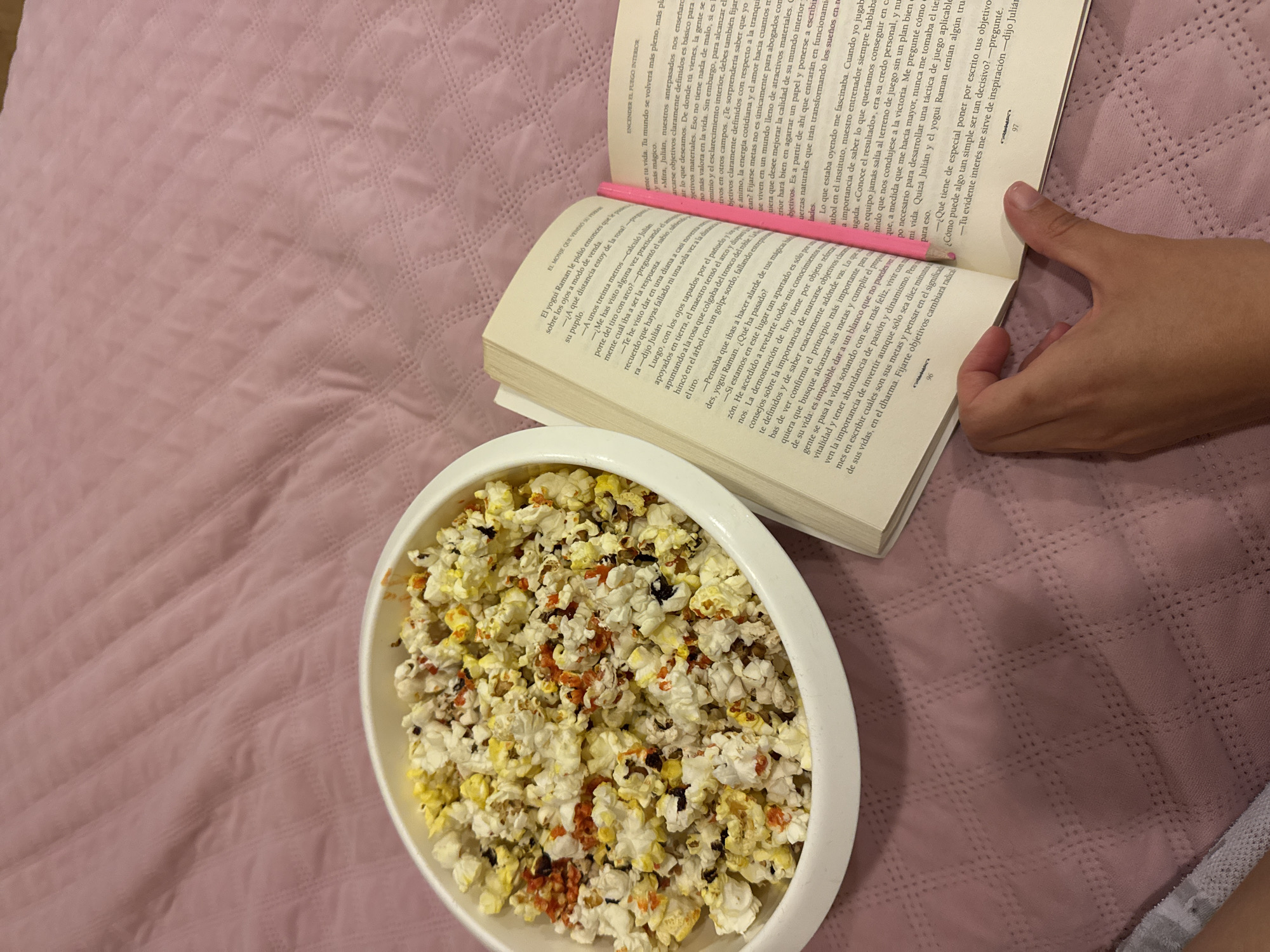 A photograph shows a book lying open on a bed next to a bowl of popcorn. A hand is holding the book open, with a pencil being used as a bookmark. The book appears to be in Spanish, with two pages visible and text on each. The bowl, filled with popcorn that includes some reddish seasoning, sits on the bed next to the book. The bed has a textured, patterned pink bedspread. The setting appears to be indoors, perhaps in a bedroom or living space. Based on the text visible in the book, it does not appear to be in Tlaxcalancingo, Mexico. It is a scene of someone reading and snacking, likely in a relaxed, personal setting.