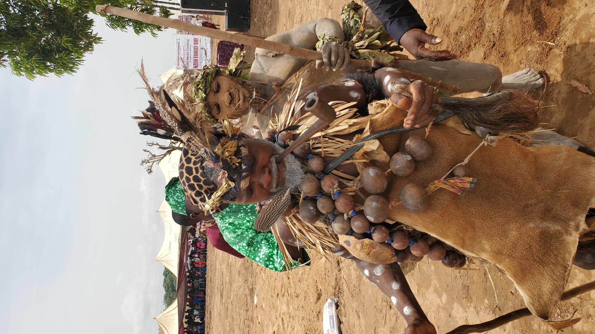 The image depicts two individuals participating in a cultural event, likely in a public open space in the city of Akwanga, Nigeria. The foreground figure wears a elaborate headdress, facial paint, and a garment adorned with spherical objects. This person carries what appears to be an animal hide. Behind and above this figure is a second individual covered in what appears to be mud or clay and also wearing foliage. This person is holding a wooden pole. The background shows tents and a crowd of people, along with a sign that mentions "Nzeh Mada Festival" and "NZEH FAMILY DAY".