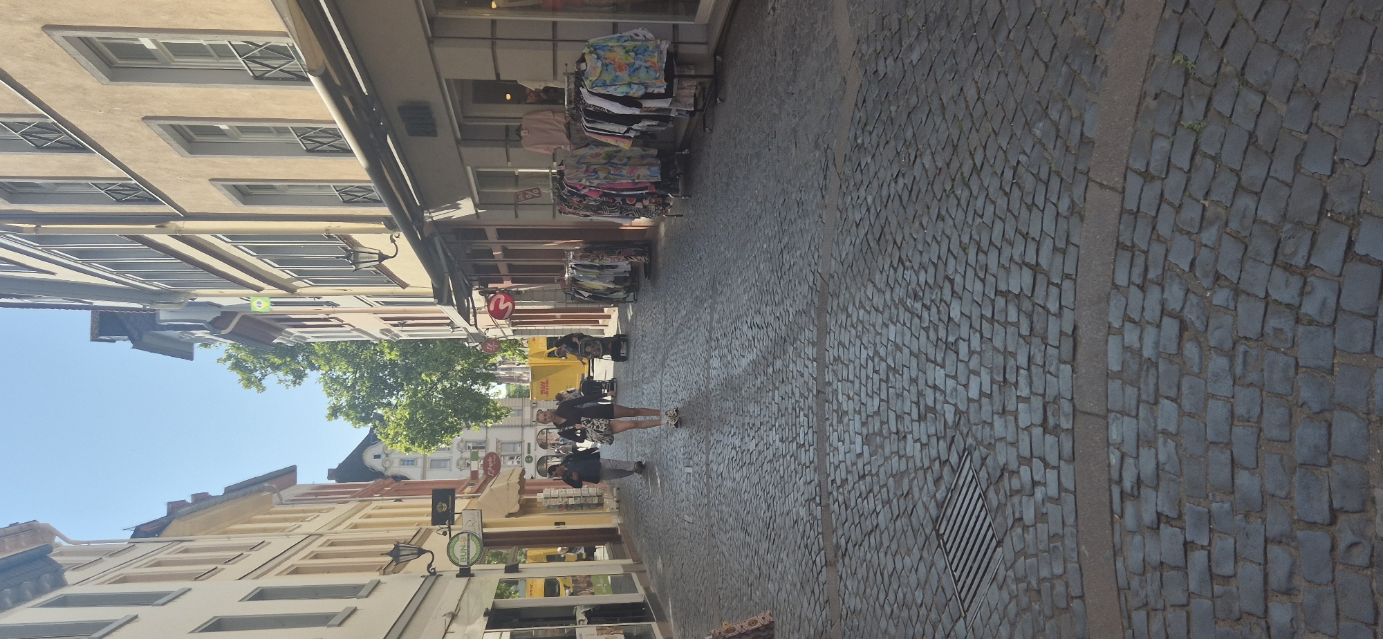 The image shows a narrow cobblestone street in Mainz, Germany, lined with traditional European buildings. A few people are walking along the street. In the foreground, the cobblestone pavement is detailed, with a metal grate on the left. Several shops with awnings are visible on both sides of the street. Clothes are displayed on racks outside some of the shops. A yellow DHL truck is parked further down the street. The sky is clear and blue, indicating a sunny day.