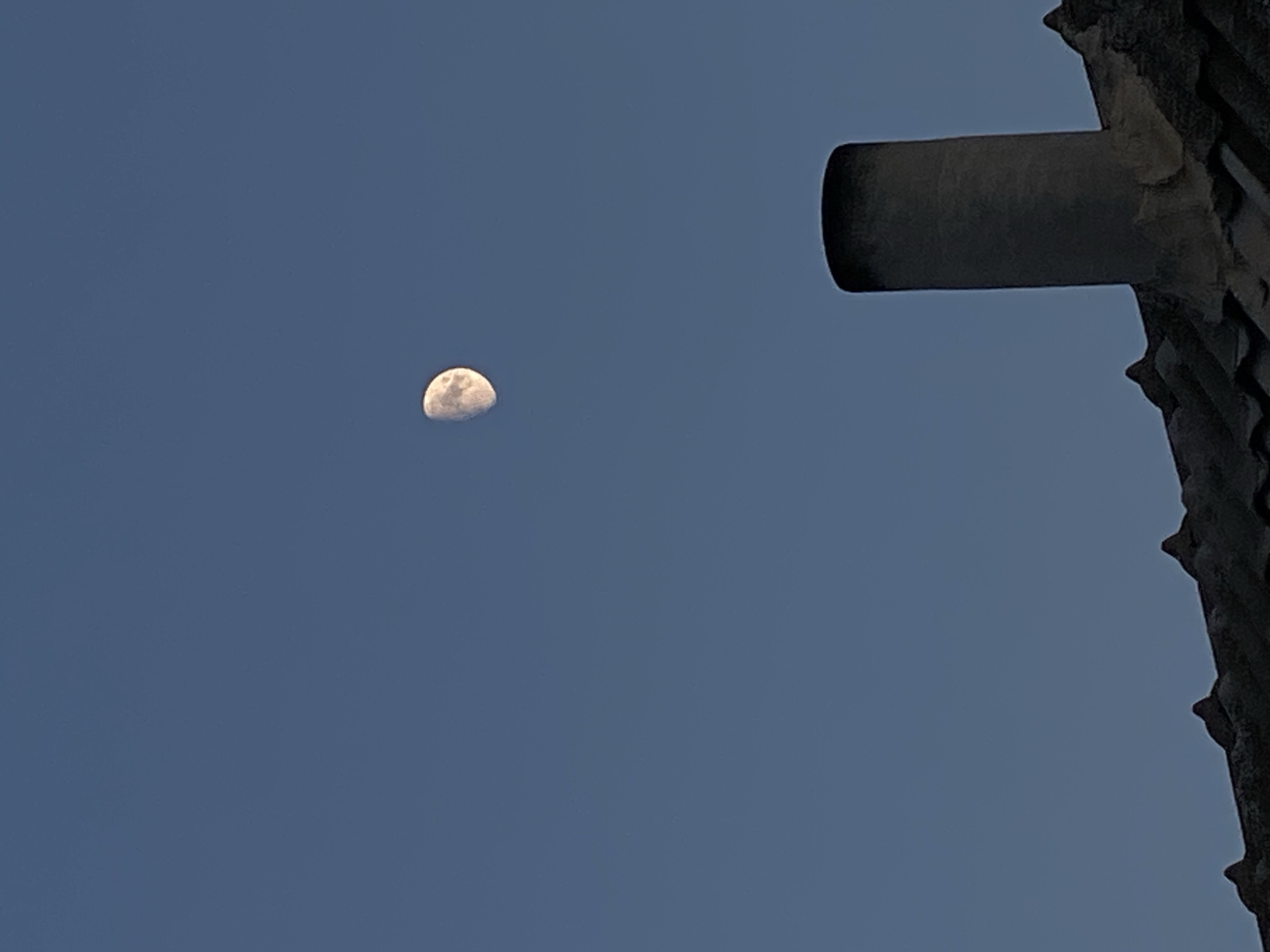 The image shows a partially illuminated moon in a clear blue sky. In the foreground, the roof of a building is visible, with a chimney standing tall. The roof tiles are dark and weathered, and the chimney is cylindrical and appears to be made of concrete. The overall scene suggests an outdoor setting, possibly in a residential area. The specific location of Cairaclia, Moldova, is provided as context for the image.