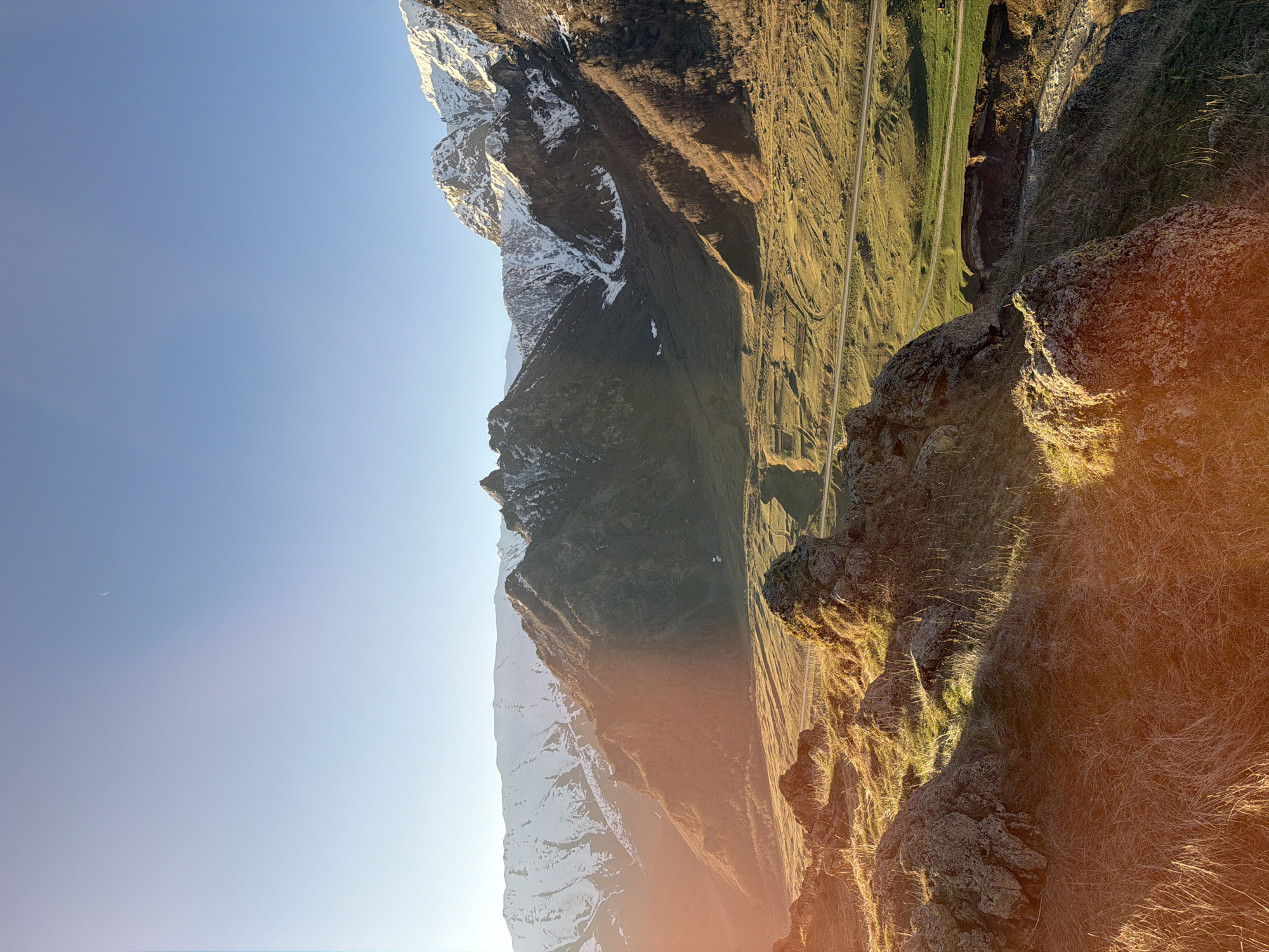 The image shows a mountainous landscape. In the distance, snow-capped peaks are visible. The mid-ground features steep, rocky terrain with patches of green vegetation and a road. The foreground consists of rocks and dry grass. The sky is clear and blue. There is a lens flare in the lower right corner. The location is possibly near Pkhelshe, Georgia, based on the surrounding environment. There are no people visible in the image.