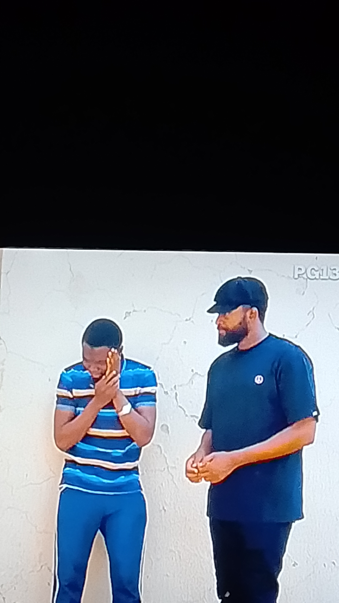 The image shows two men standing in front of a textured, off-white wall. The man on the left is wearing a blue and white striped polo shirt with yellow accents, and blue track pants with white stripes. He has his hands clasped to his face, appearing to be distressed or contemplative. A white watch is visible on his left wrist. The man on the right is wearing a dark blue t-shirt with a small white logo and a black baseball cap. He has a beard and is looking to the left. His hands are clasped in front of him. The background wall appears to be outdoors, and in the upper right corner, text that appears to read "PG13" is visible. The lighting suggests it is daytime. This scene is likely located in Lokoja, Nigeria, given the context of the provided image.