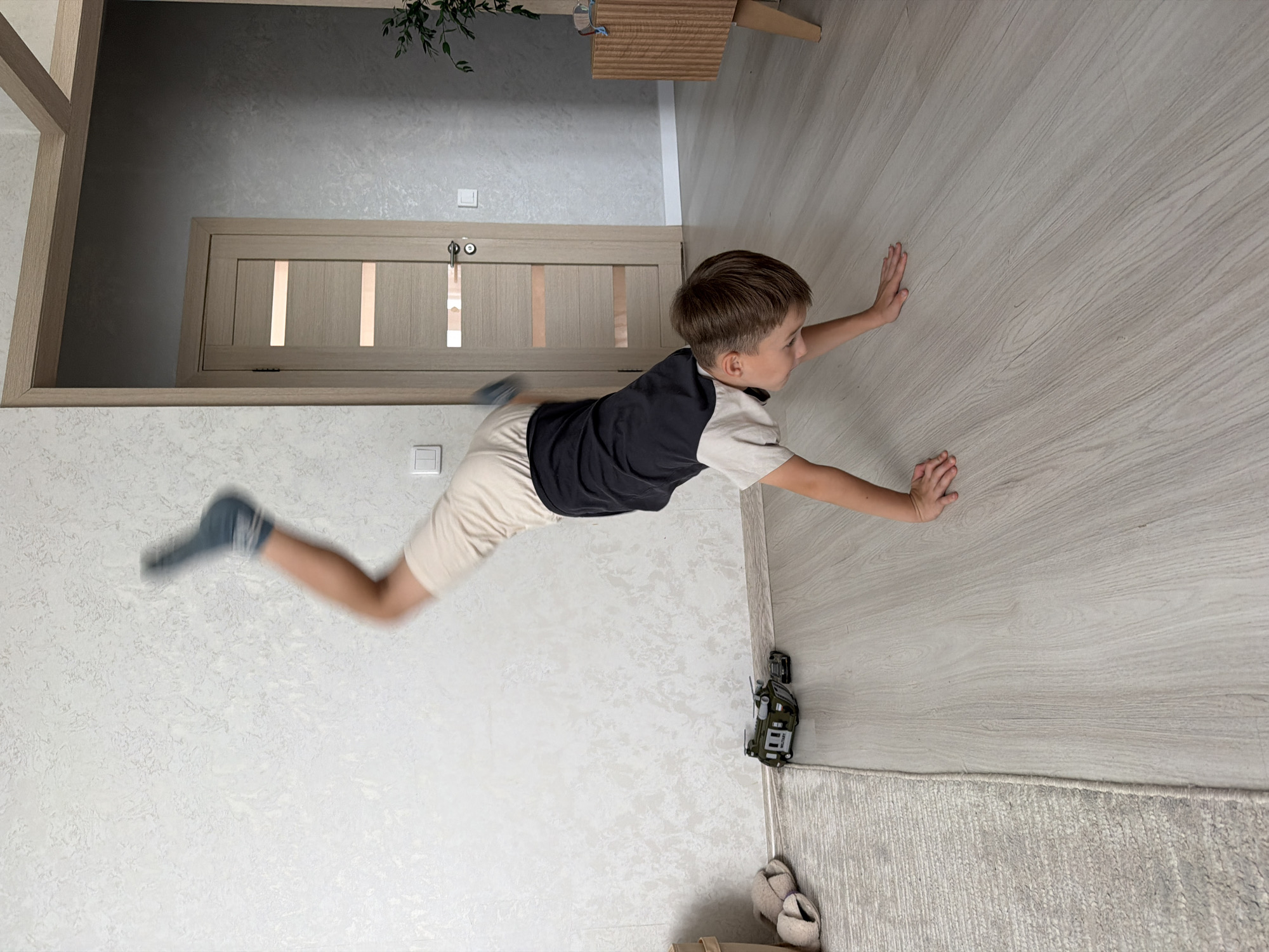 A young boy is captured mid-air, in a dynamic pose that suggests he is jumping or falling. He is wearing a dark t-shirt with a white collar and light-colored shorts, along with dark socks. His arms are outstretched, and his legs are bent. The boy appears to be in an indoor setting, possibly a hallway or living room, with light-colored walls and a wooden floor.
The background shows a closed wooden door with vertical panels and a metallic handle. A plant is visible in the upper part of the frame, and a wooden shelf or cabinet is partially seen. In the lower right corner, a small toy vehicle is placed on the floor near a rug and a pair of slippers. The lighting suggests it's daytime, and there are no indicators of weather conditions. The boy's expression is not clearly visible due to his movement and the angle, but the action itself conveys a sense of playful energy or excitement. The location is identified as Kazan, Russia. There is no visible text in the image.