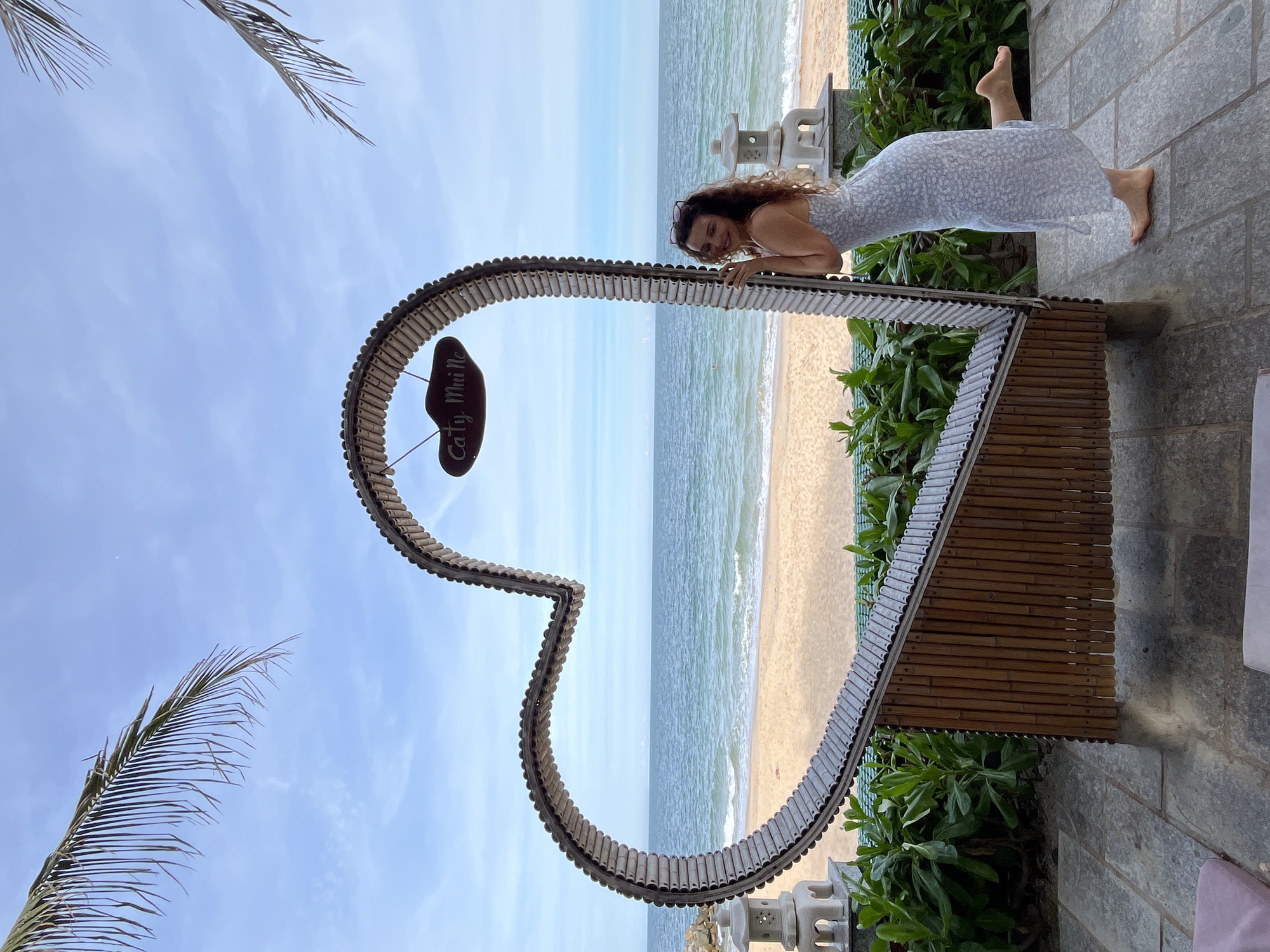A woman with curly brown hair and glasses perched on her head, wearing a light blue floral print dress, stands next to a large, hollow heart-shaped structure on a paved patio. The structure is made of bamboo and what appears to be small, segmented metallic pieces. A sign reading "Caty Mui Ne" hangs from the top of the heart. The woman is leaning on the heart, with her right leg extended back and her left foot on the ground. Behind her, a sandy beach meets the blue ocean under a cloudy sky, with palm fronds visible in the upper corners of the frame. Two stone lanterns are positioned on either side of the heart, and lush green plants grow in front of the beach. The overall scene suggests a resort or tourist area in Mũi Né, Vietnam.