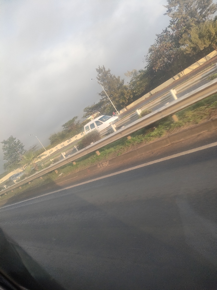 The image depicts an exterior view of a highway or major road in Theta ward, Kenya, captured from a moving vehicle's interior. The perspective is angled slightly upwards from the lower left.
In the immediate foreground, dark asphalt pavement is visible, with a solid white line marking a lane edge. White numbers "4130" are stenciled on the asphalt surface near a metallic guardrail.
A silver-colored, four-bar guardrail extends horizontally across the midground, separating the foreground lane from an elevated or adjacent section of the road. Beyond the guardrail, a light-colored concrete barrier wall features partially legible white text. Behind this barrier, a multi-lane road is visible. A white van or SUV is traveling away from the viewer on this road. Further down, a dark sedan is partially visible. A single individual, possibly wearing a red top, is situated on the far side of the road near the white van, either standing or walking.
The background consists of dense green foliage from trees and shrubs lining the roadside, punctuated by tall streetlights. The sky is uniformly overcast and light grey, indicating diffuse lighting conditions.