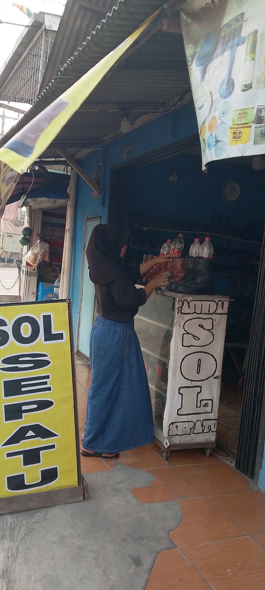A woman wearing a dark headscarf and a long blue denim skirt stands at a storefront. She is facing away from the camera, reaching towards a display case containing boots and several plastic bottles of liquid. To the left of the woman is a large yellow sign with black lettering that appears to read "SOL SEPATU", suggesting the establishment sells shoe soles or shoes. In the background, the storefront has blue walls and an open entrance. The floor is tiled with terracotta-colored tiles and patches of concrete. The scene appears to be outdoors, with a corrugated metal roof extending over the storefront. The location is identified as Satriamekar, Indonesia.