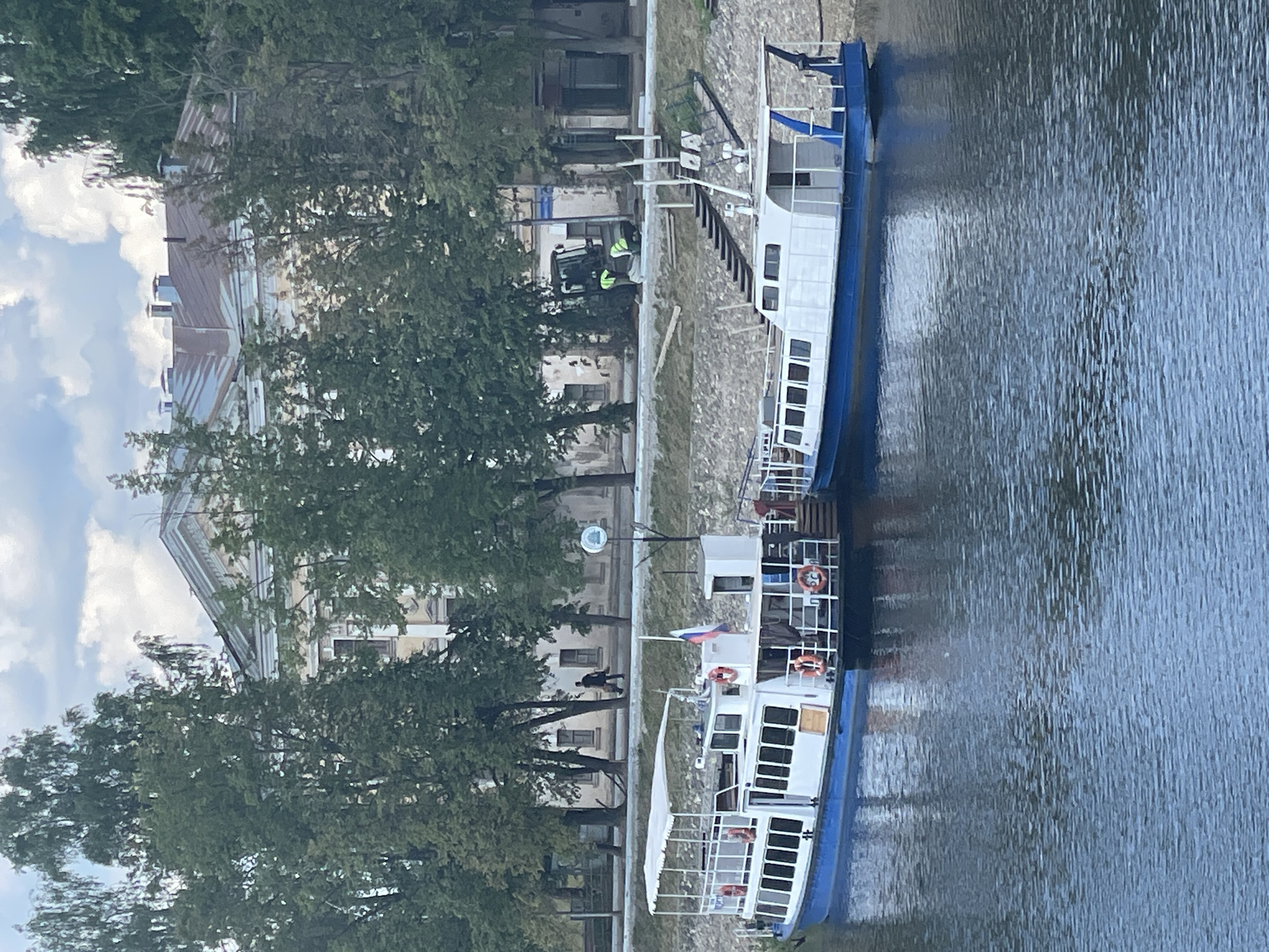 Two white and blue passenger boats are docked on a calm body of water. The boat on the left has a white canopy over its deck, multiple windows along its side, and life preservers attached to its railing. A Russian flag is visible on its stern. The boat on the right is similar in design, with windows and a blue railing. In the background, behind a stone embankment and trees, is a large, light-colored building with a gray, gabled roof. A person in dark clothing is walking on a path near the building, and a tractor with two people in high-visibility vests is visible on the embankment. This scene is located on the Vologda River in Vologda, Russia, with the prominent building likely being part of the historic Vologda Kremlin or a nearby administrative building. The sky is partly cloudy.