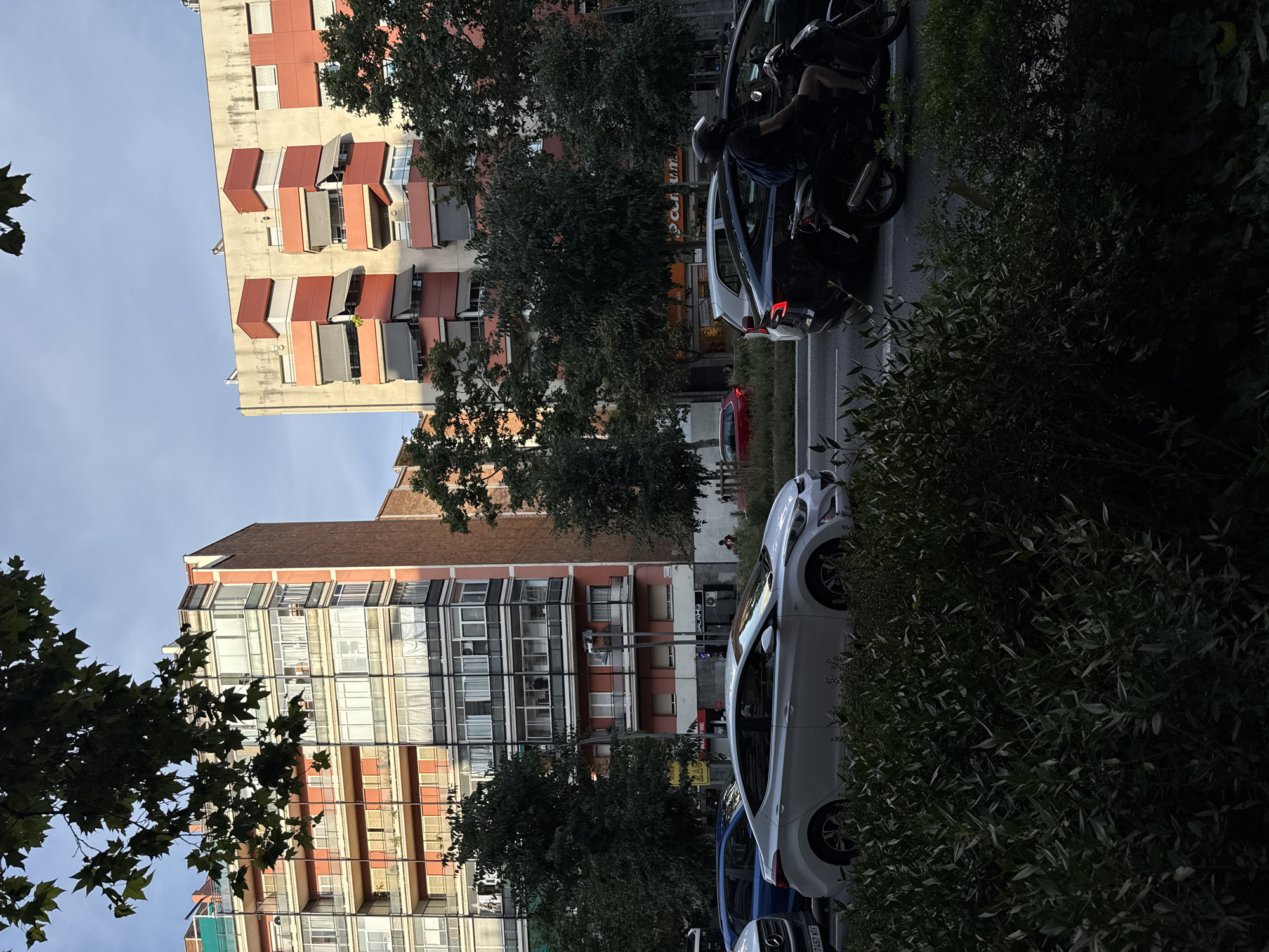 This image depicts a street scene in Barcelona, Spain, likely during the daytime, given the lighting conditions. In the foreground, a dense hedge of dark green leaves partially obstructs the view of the street. Beyond the hedge, a white sedan is parked on the right side of the road. To its left, a black SUV is visible, and a person on a motorcycle with a helmet is riding in the right lane. The street appears to be a multi-lane road with traffic.
Behind the street, two multi-story buildings are prominent. The building on the left is a salmon-pink color with multiple balconies and large windows. The building on the right features a lighter facade with red and orange elements, including awnings extended over some windows. Trees with dark green foliage are interspersed between the buildings and in front of them, adding a natural element to the urban landscape. The sky above is a pale blue with some wispy clouds, suggesting fair weather. The architecture of the buildings, particularly the balconies and the style of the awnings, suggests a Mediterranean or European urban setting. The presence of a motorcycle and cars indicates active city life.